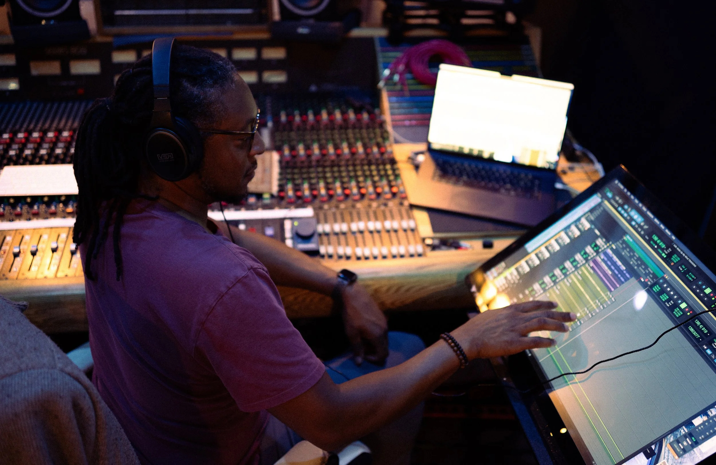 Music producer wearing headphones working on a digital audio workstation with mixing console and laptop in a recording studio.