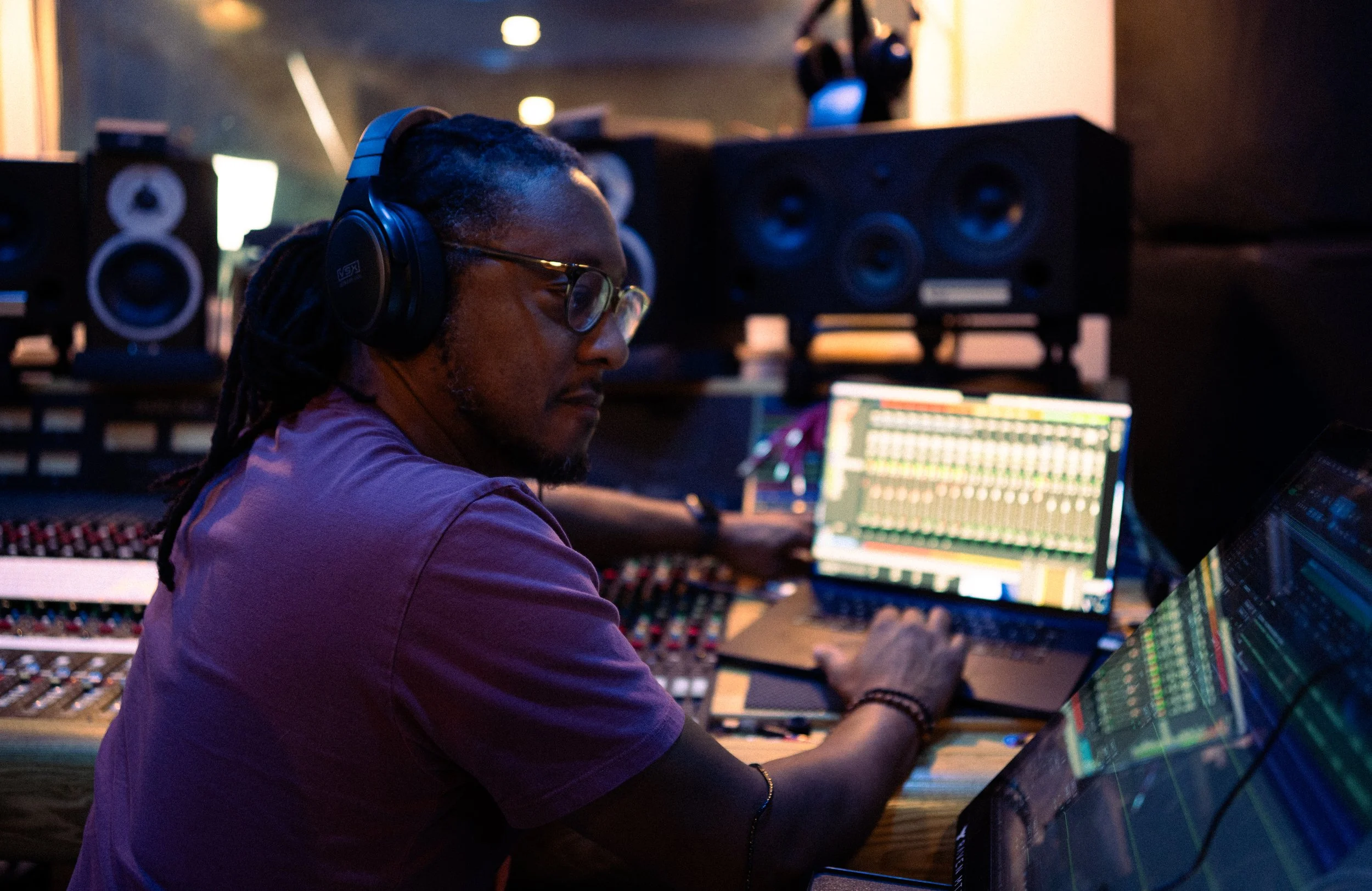 A man with dreadlocks, glasses, and headphones working at a music production studio with a computer displaying a digital audio workstation.