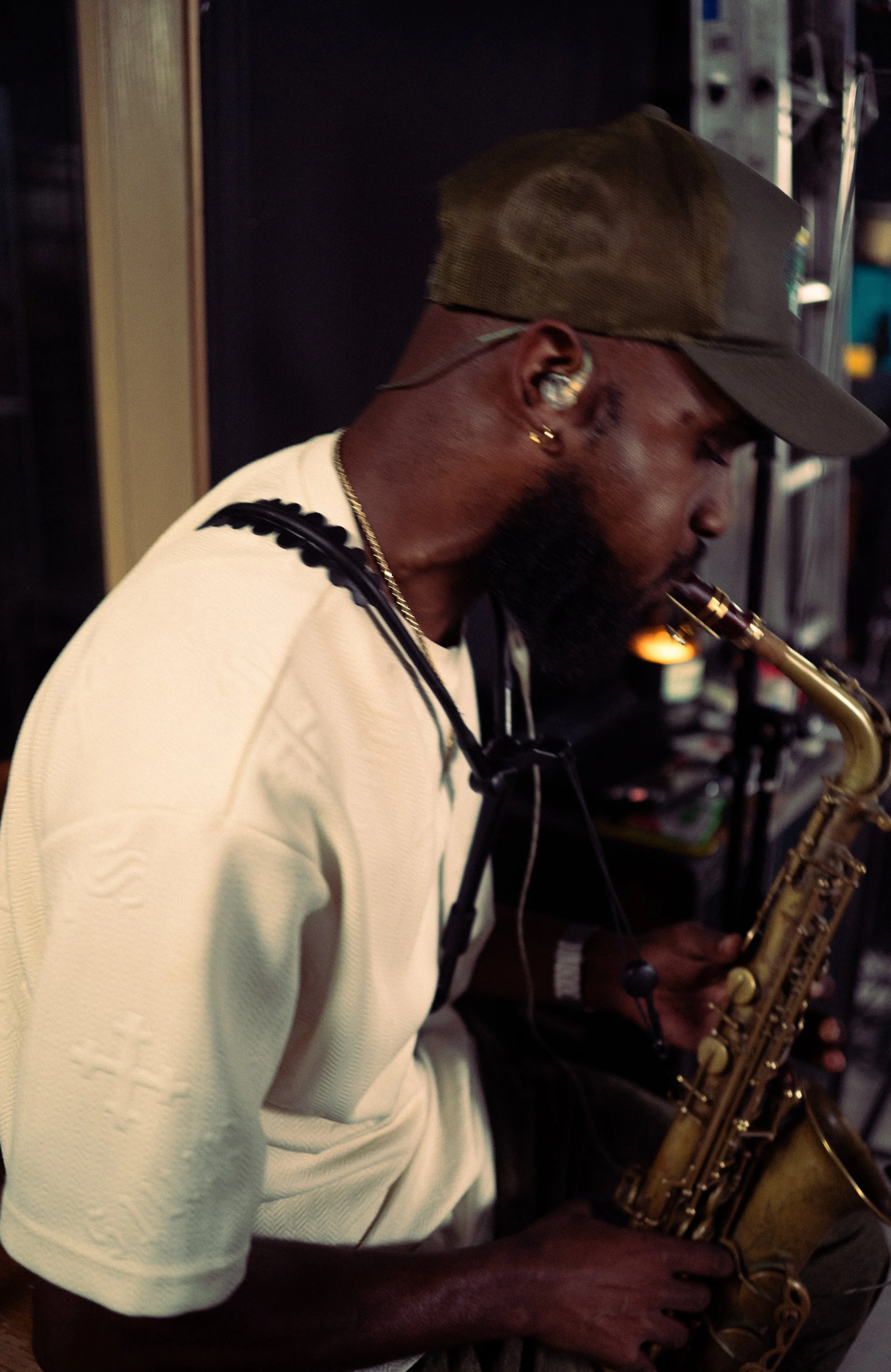 A man wearing a baseball cap, earrings, and a white shirt plays a saxophone, with headphones around his neck, in an indoor setting.