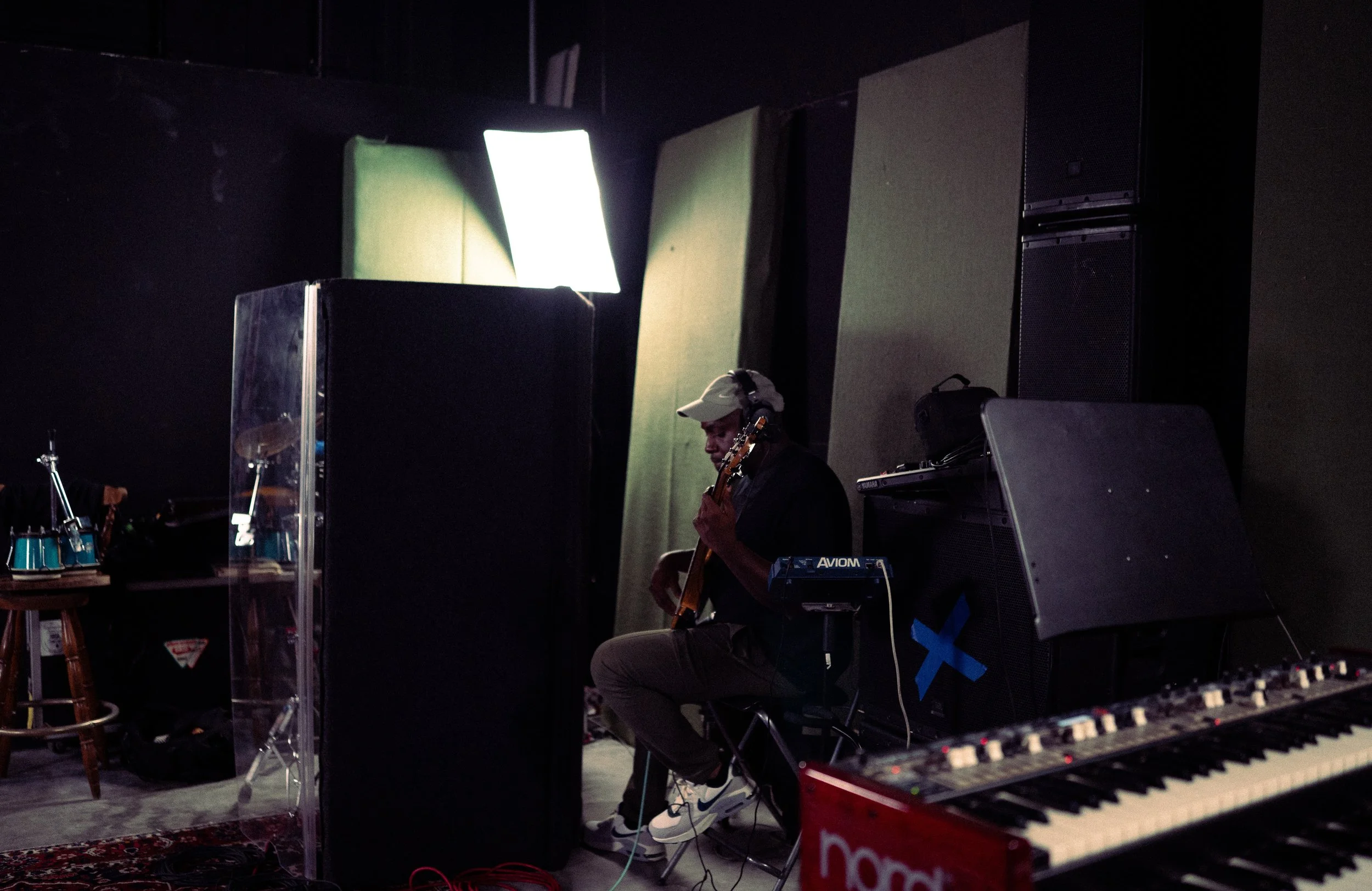 A person wearing a white cap and headphones is sitting in a music studio, playing an electric guitar surrounded by various music equipment, including a keyboard and speakers.