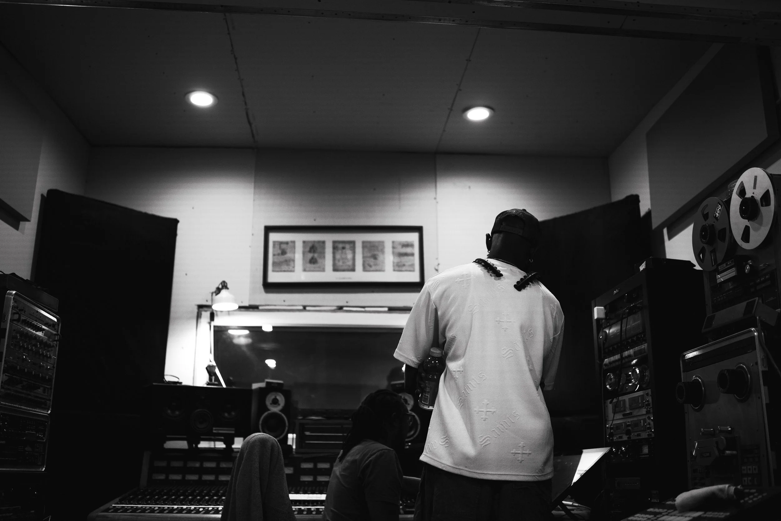 Two people working in a music recording studio surrounded by audio equipment. One person stands with their back to the camera, holding a bottle, while the other is seated at a mixing console.