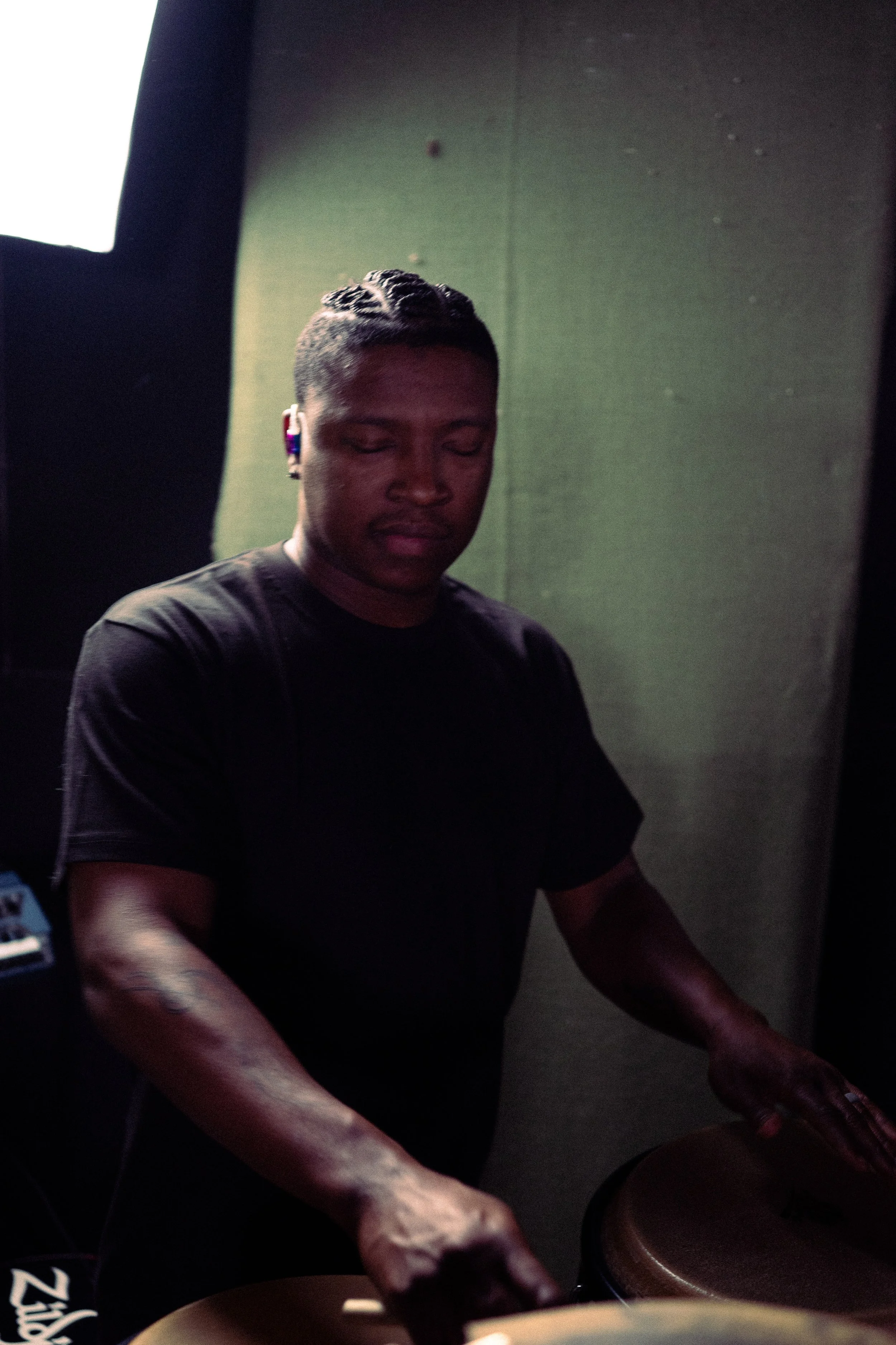 A young man DJing in a dark room, wearing a black t-shirt, with tattoos on his arm, focused on the turntables, and wearing wireless earbuds.