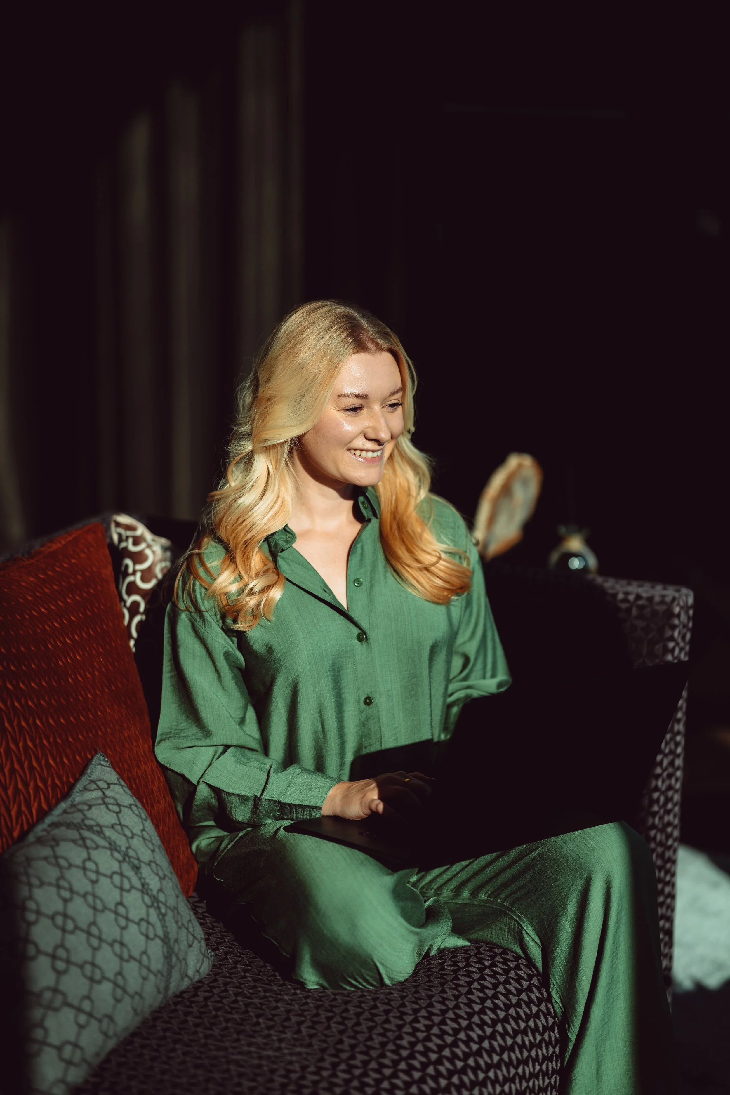 Woman with long blonde hair sitting on a patterned sofa, using a laptop in a dimly lit room.