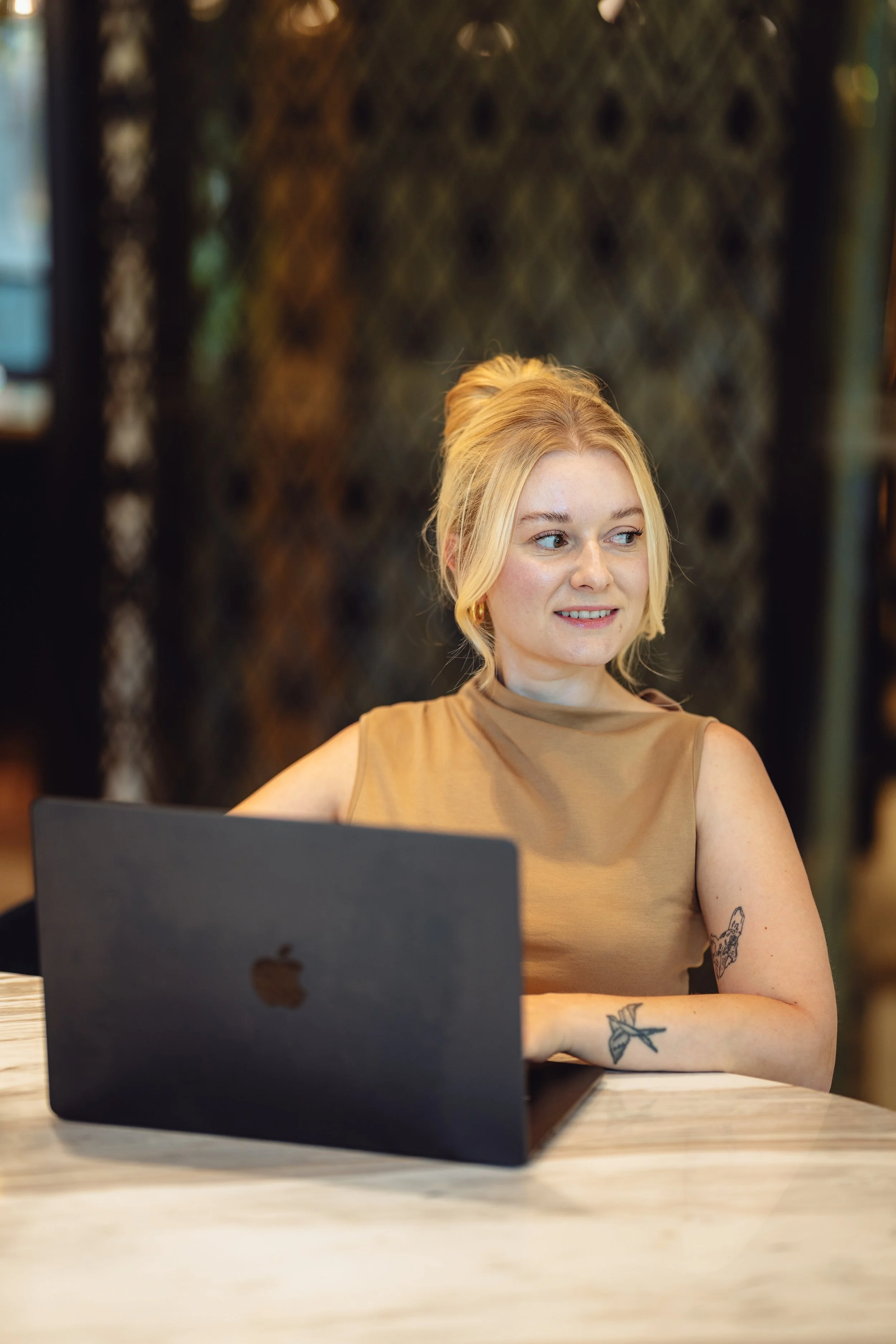 A young woman with blonde hair tied in a bun, wearing a tan sleeveless top, sitting at a table with a black MacBook in a modern cafe or restaurant.