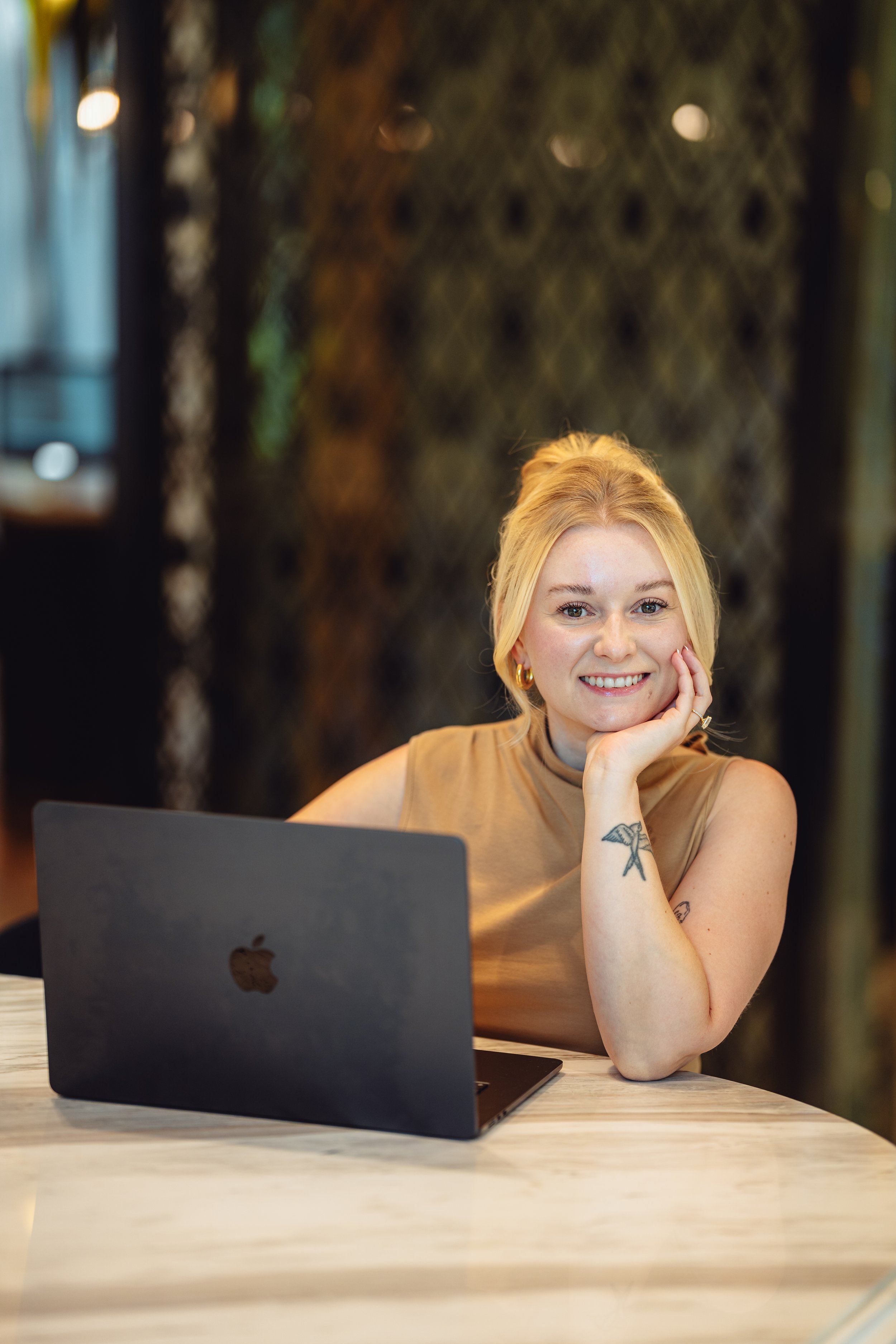 A young woman with blonde hair, wearing a sleeveless tan top, sitting at a table with a black Apple MacBook open in front of her, smiling and resting her chin on her hand in a warmly lit, stylish indoor setting.