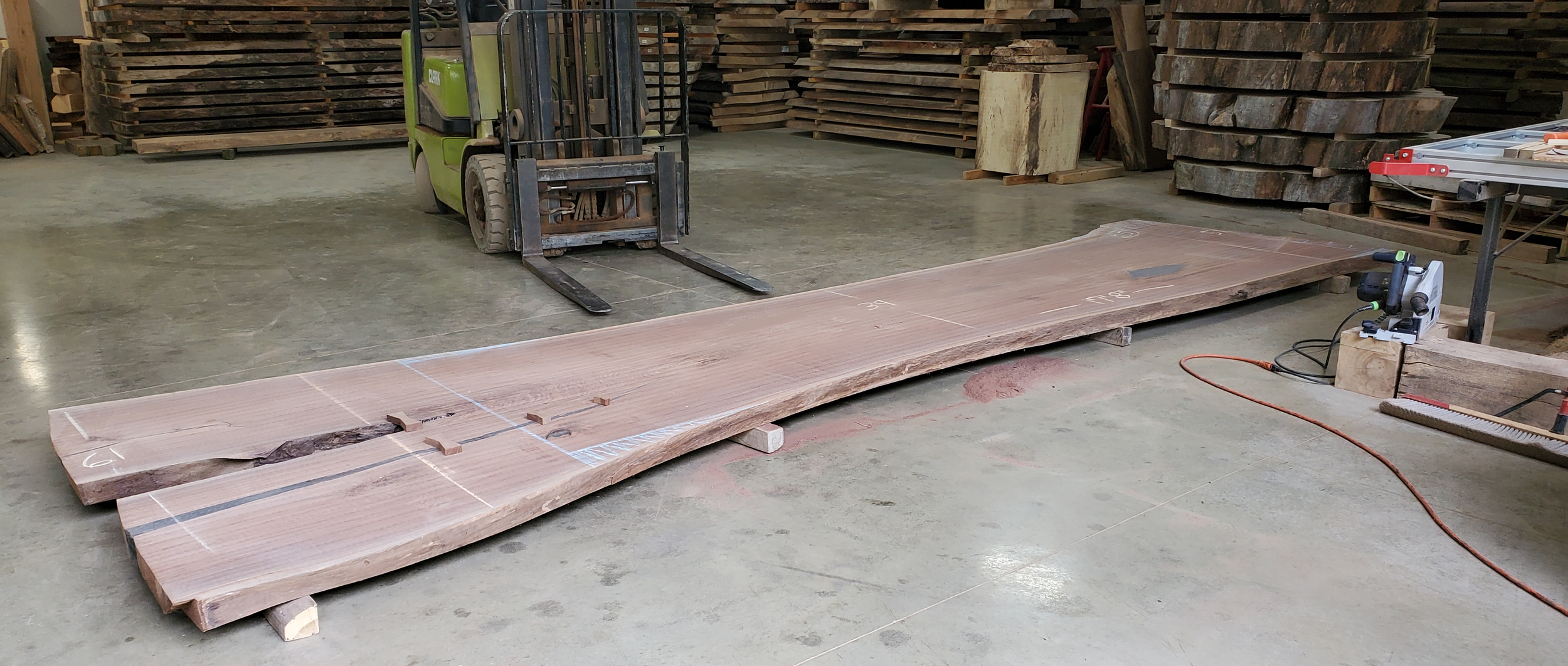 Large wooden slab on the floor in a woodworking shop, with a forklift, a table saw, and stacks of wood in the background.