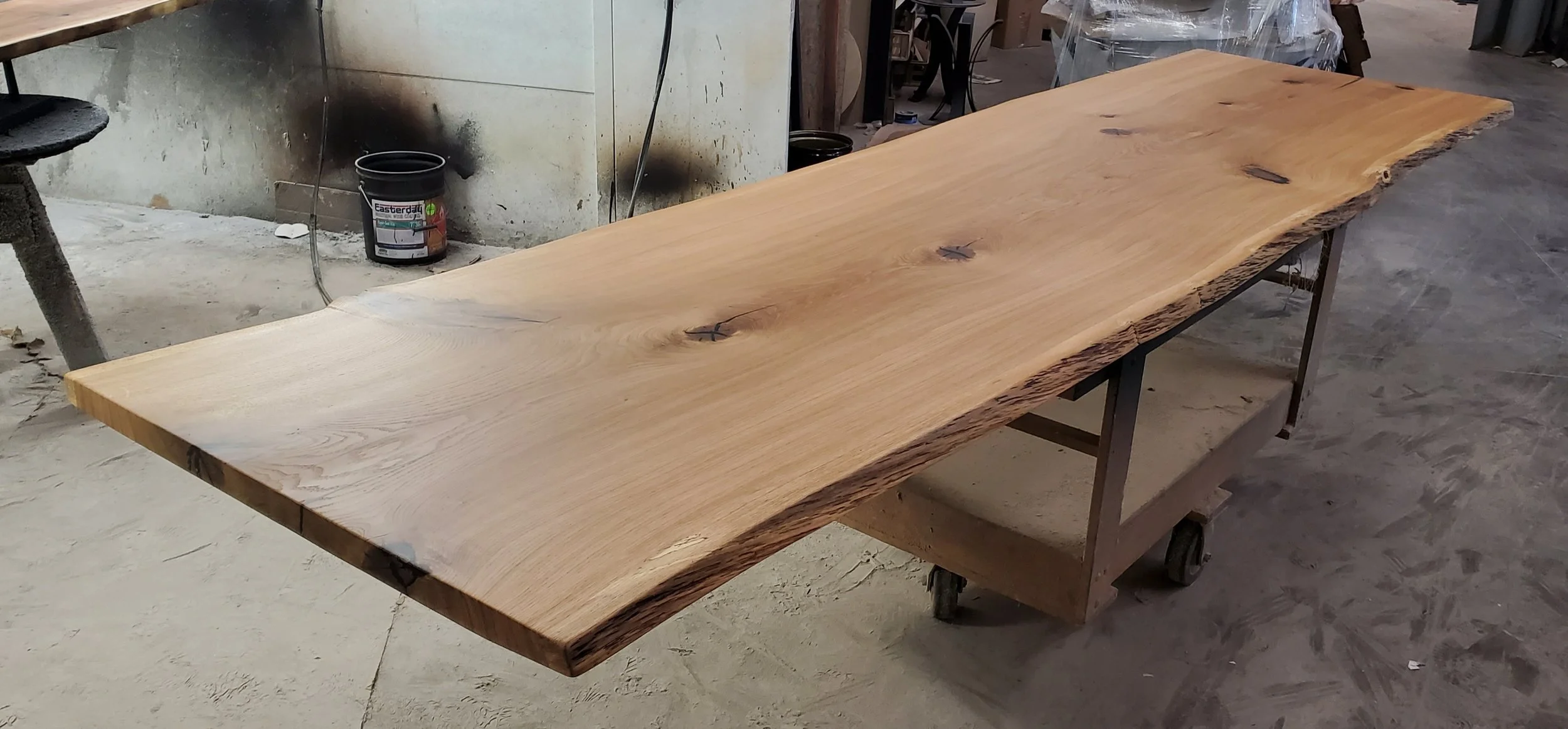 A large, unfinished wooden table with a natural, live edge design in a woodworking workshop.