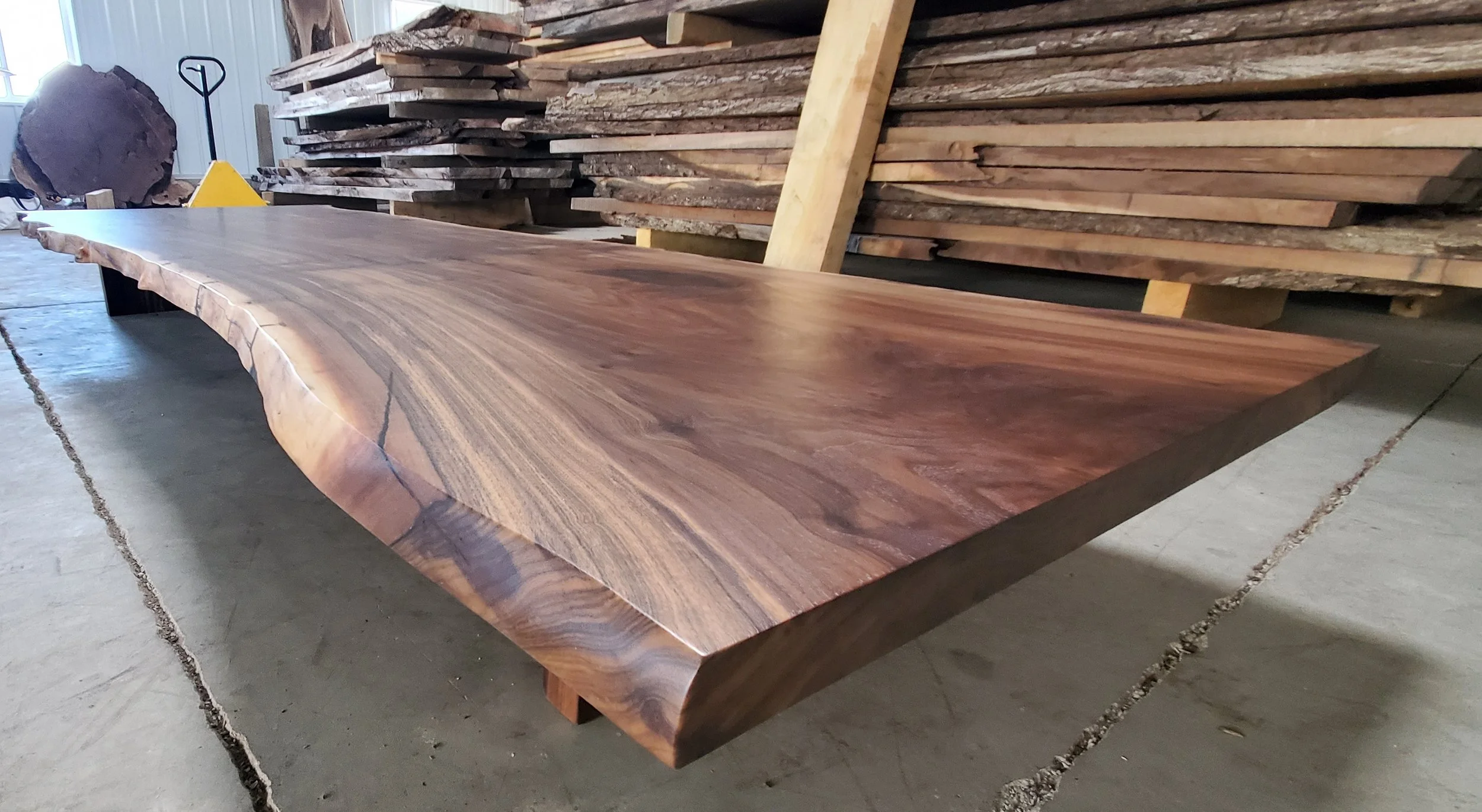 A large wooden slab with a polished surface resting on supports in a workshop, with stacked wood planks and a saw in the background.