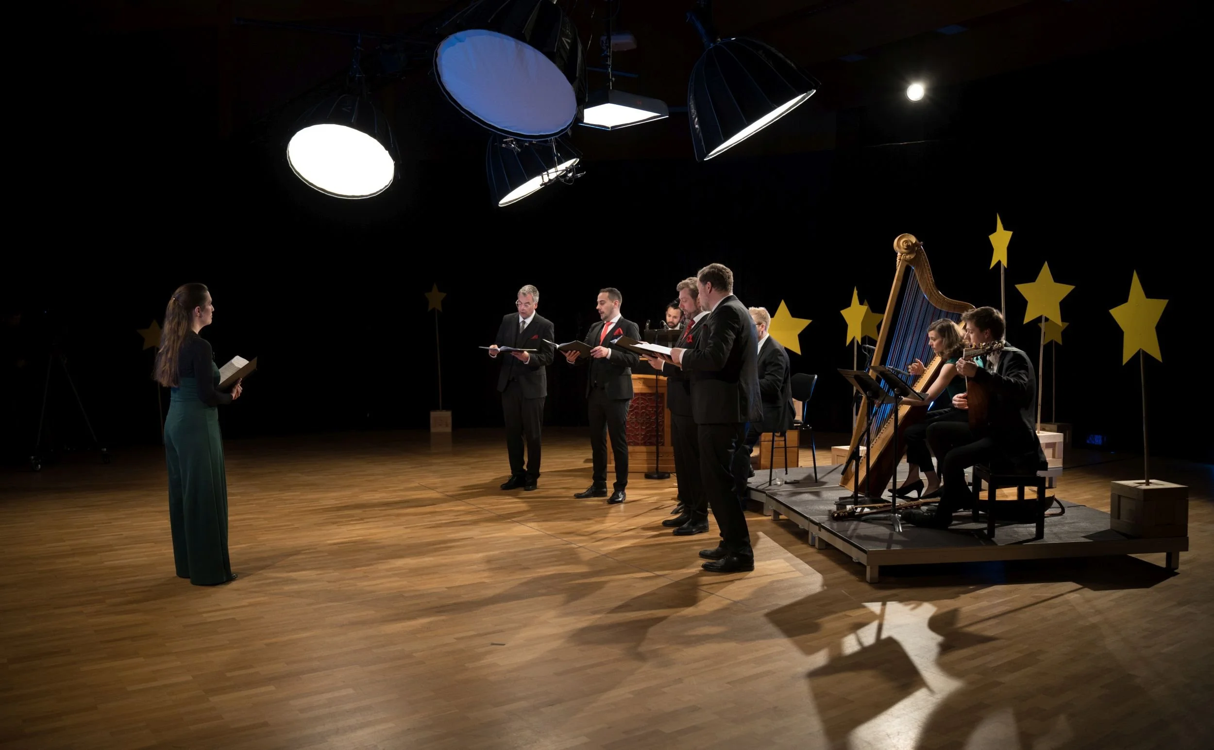 A choir rehearsal or performance with a conductor, singers, and musicians playing harp and guitar, in a decorated studio or stage with star props and bright overhead lights.