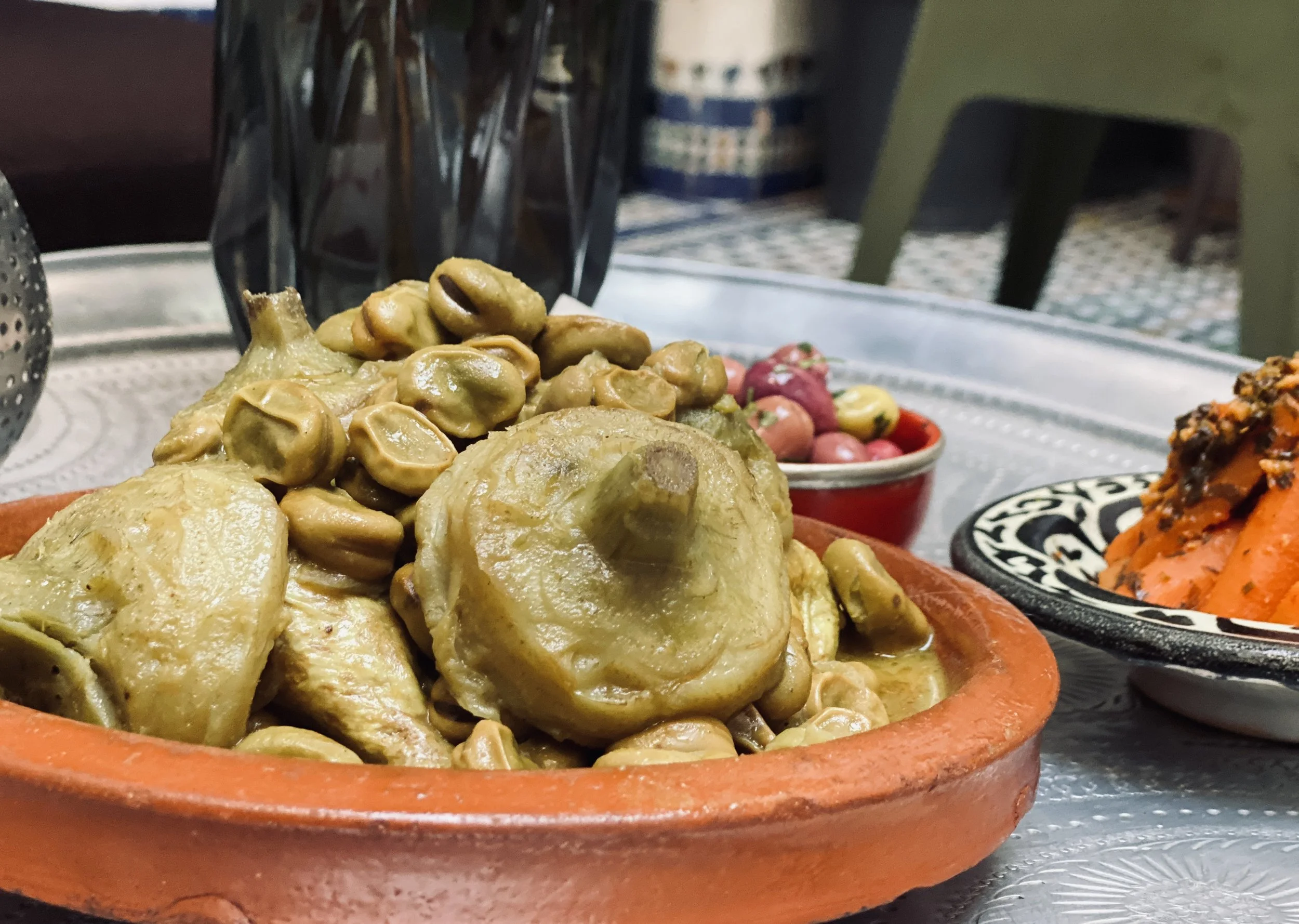 A close-up of a clay bowl filled with cooked or marinated chickpeas and chicken pieces. In the background, there is a small bowl with pink radishes, and other dishes with vibrant food, likely part of a traditional meal setup. The scene is set on a metal surface with an indoor setting.