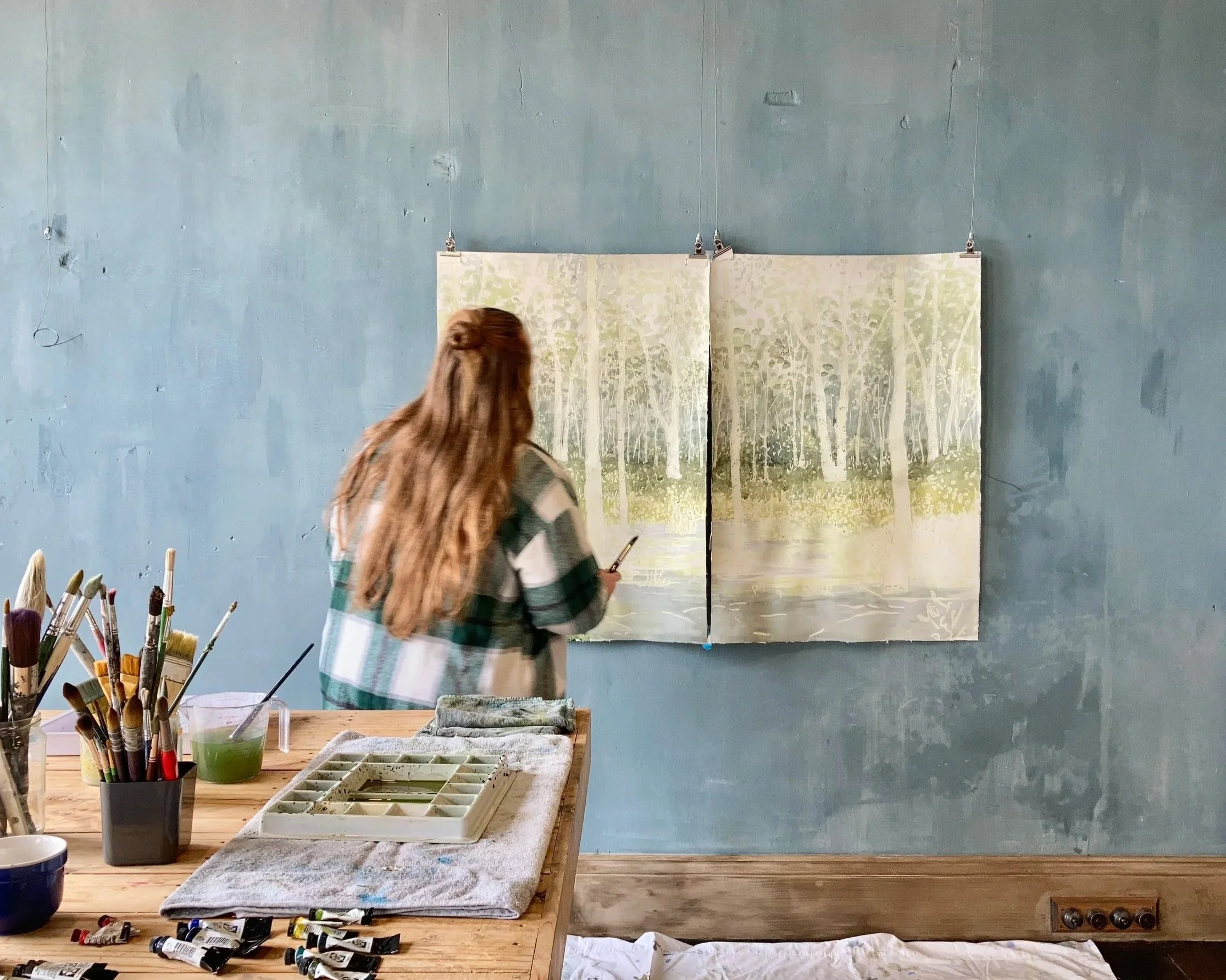 Lauren Guymer working on a painting of a forest scene, viewed from the back, with paint supplies on the table.