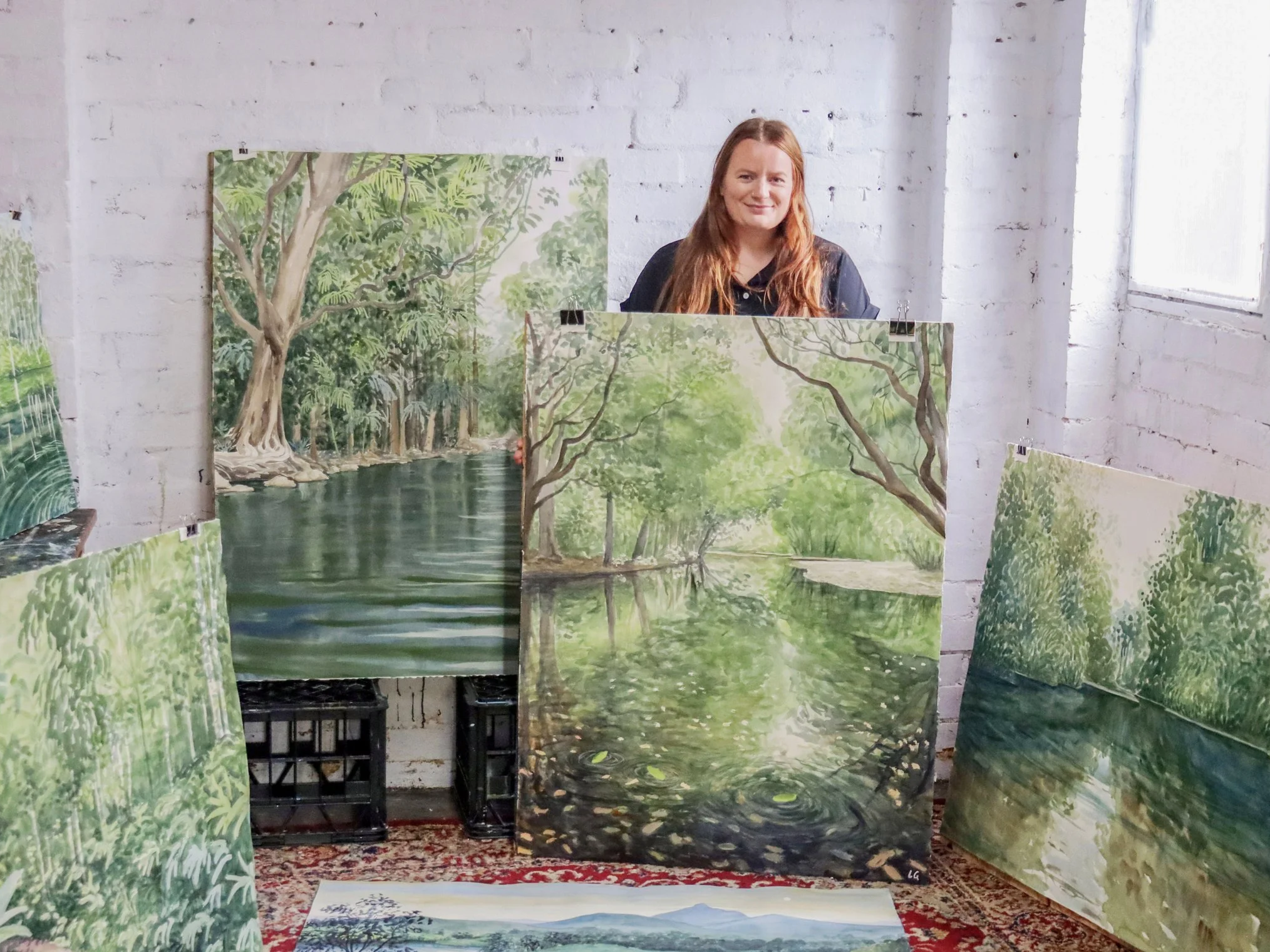A woman with long red hair standing behind five landscape paintings of rivers and trees in a white brick room with a large window.