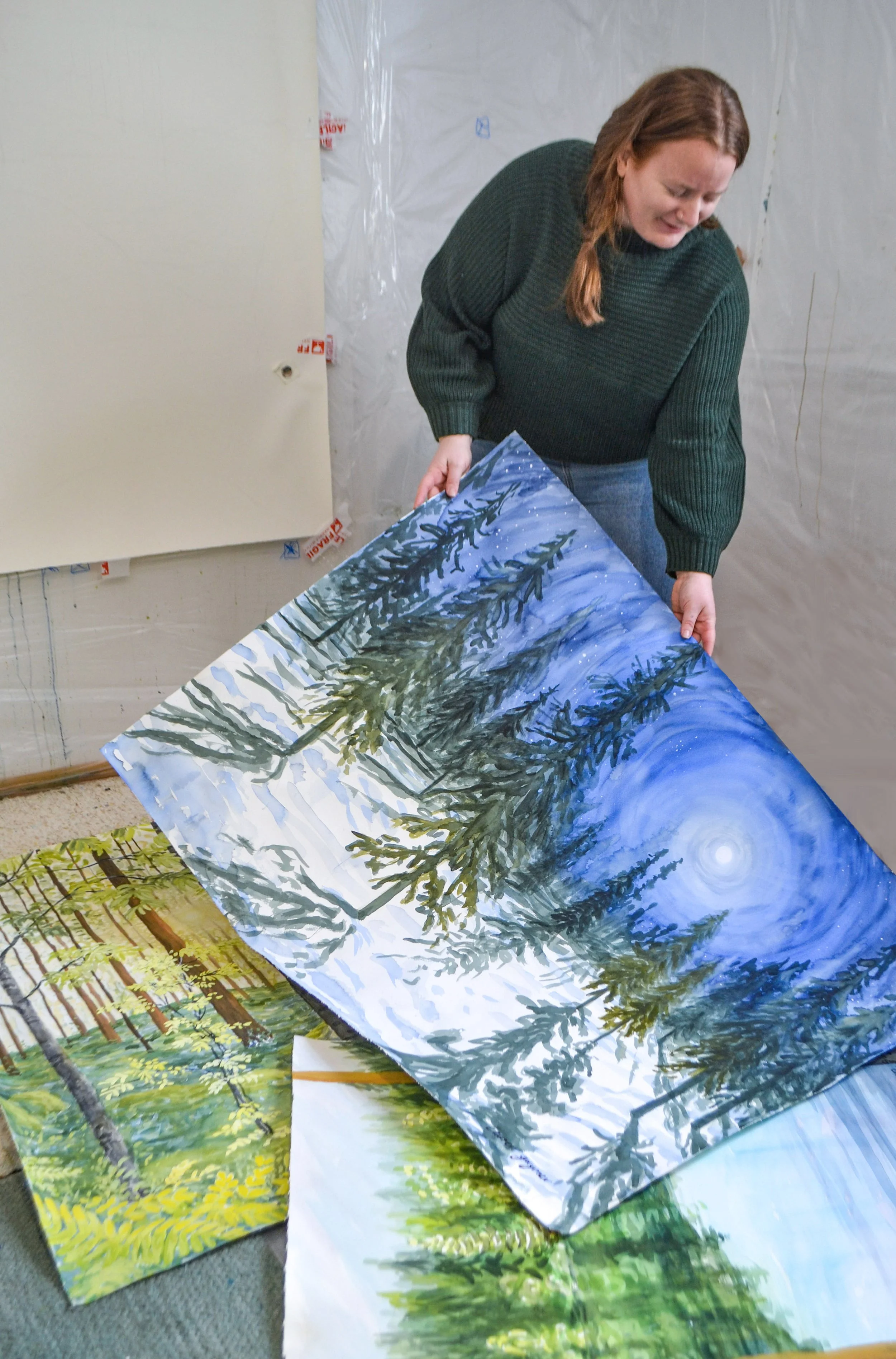 Woman holding a painting of a snowy forest scene with trees and a moonlit sky.