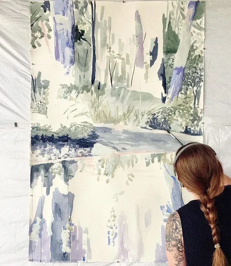 A woman with a braid and tattoo on her arm painting a landscape watercolor of a forest by a river or creek.