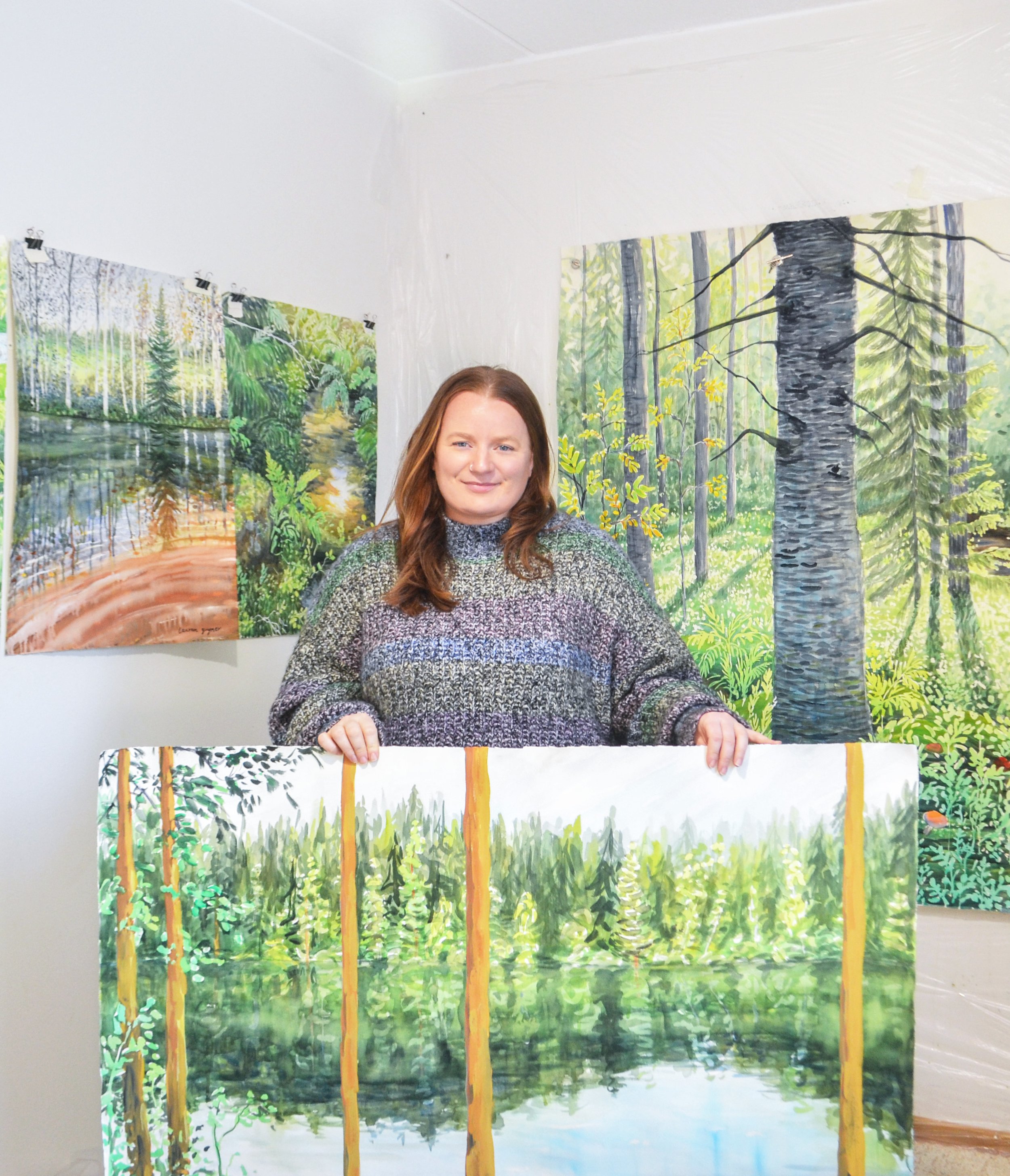 A woman holding a landscape painting of a forest and water in an art studio, with paintings of forest scenes hanging on the white wall behind her.
