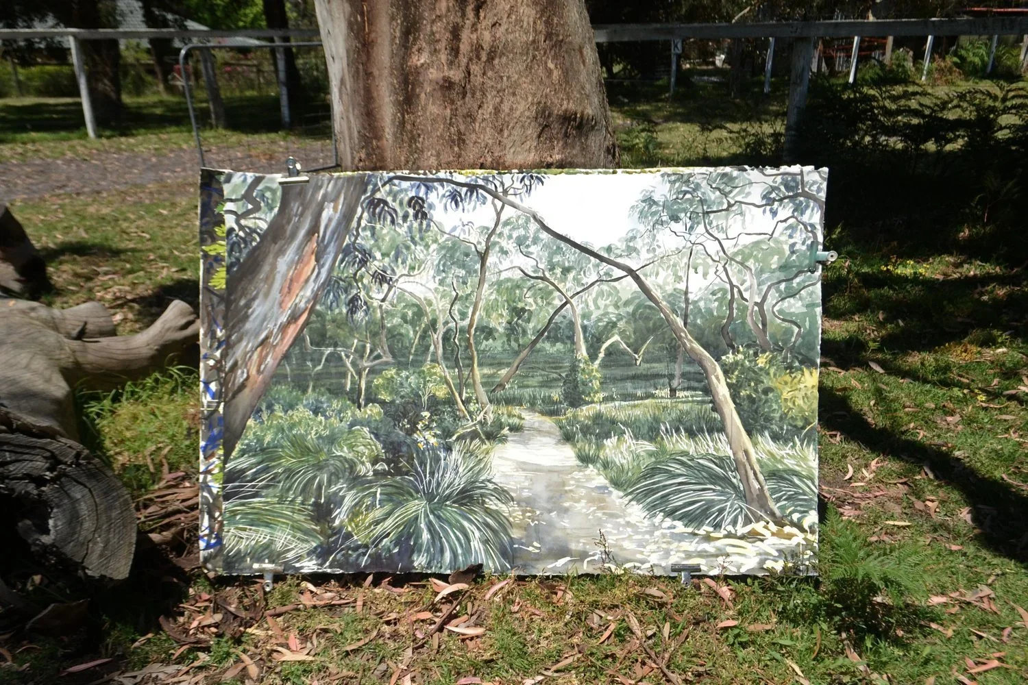 Painting of a lush forest scene with a stream, trees, and green foliage, propped against a tree outdoors.