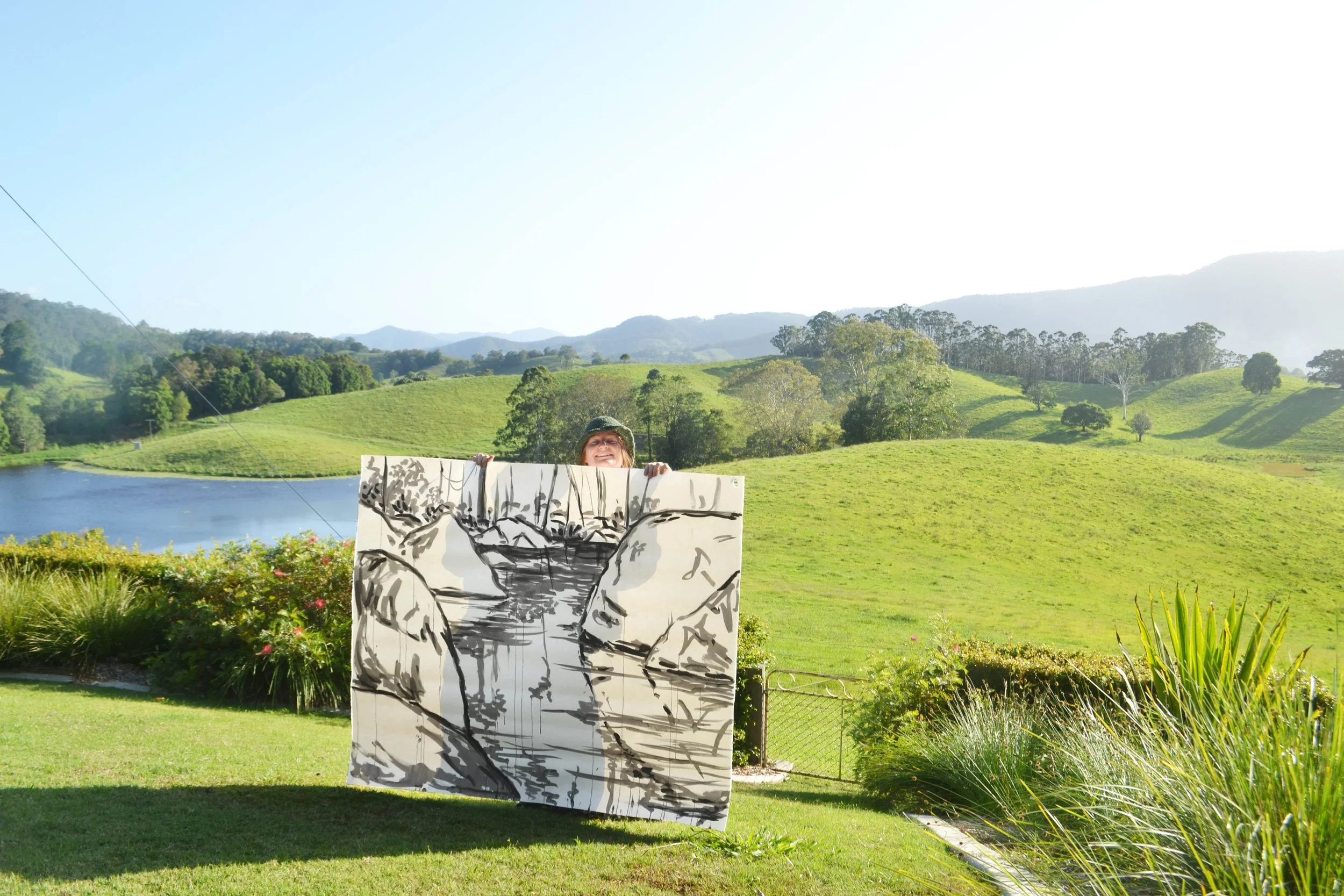 A person holding a large black-and-white painting of a mountain and river landscape outdoors near a lake and green rolling hills on a sunny day.
