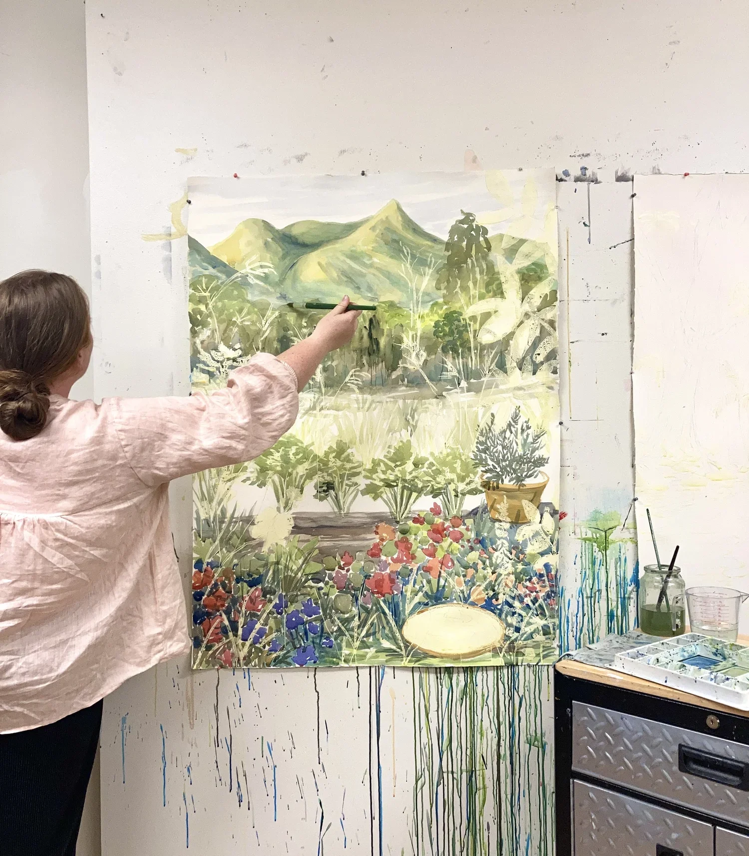 Person painting a landscape with mountains, trees, and flowers on a large canvas in an art studio.