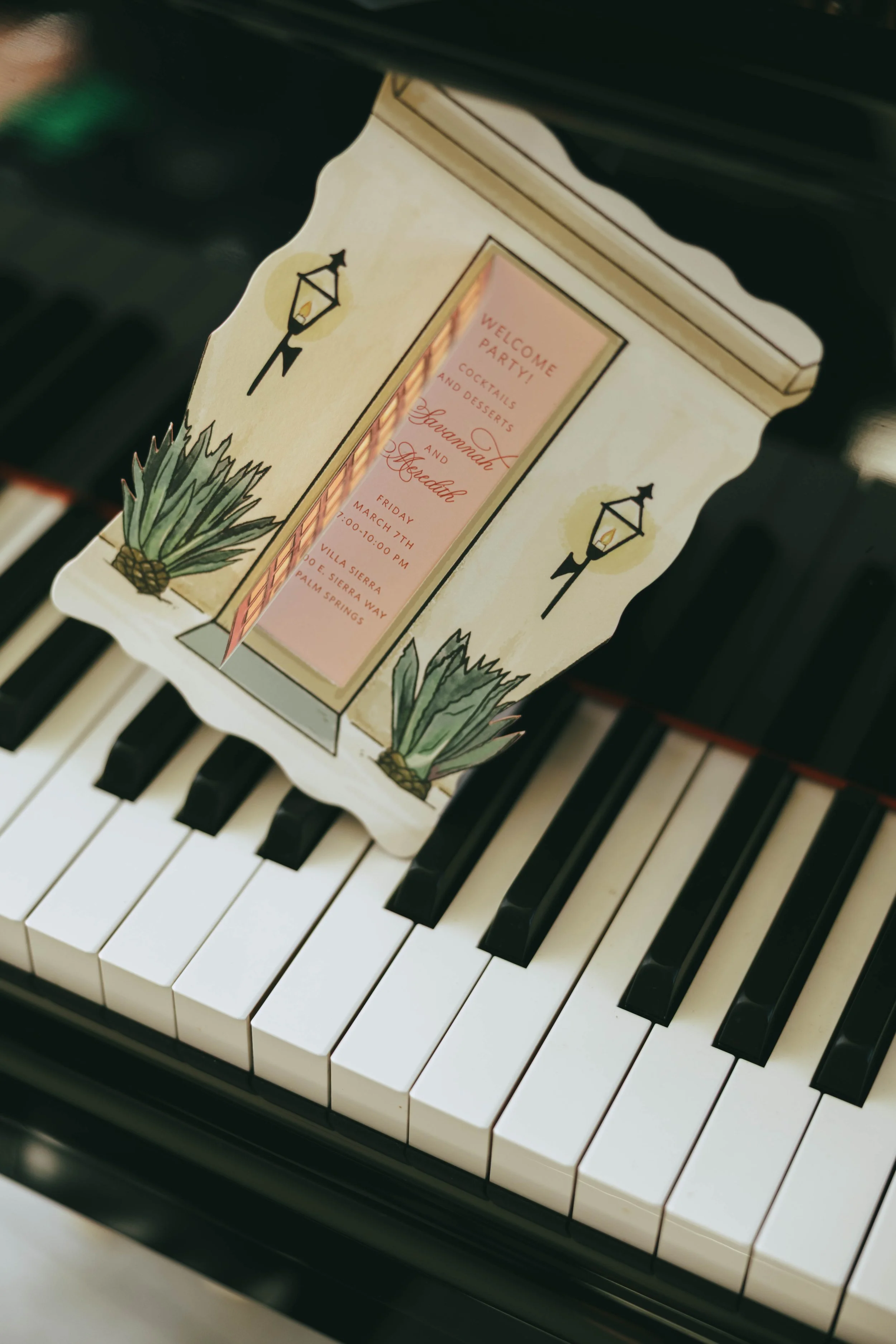 Pink and green invitation cards with cactus illustrations propped upright on the keys of a black grand piano