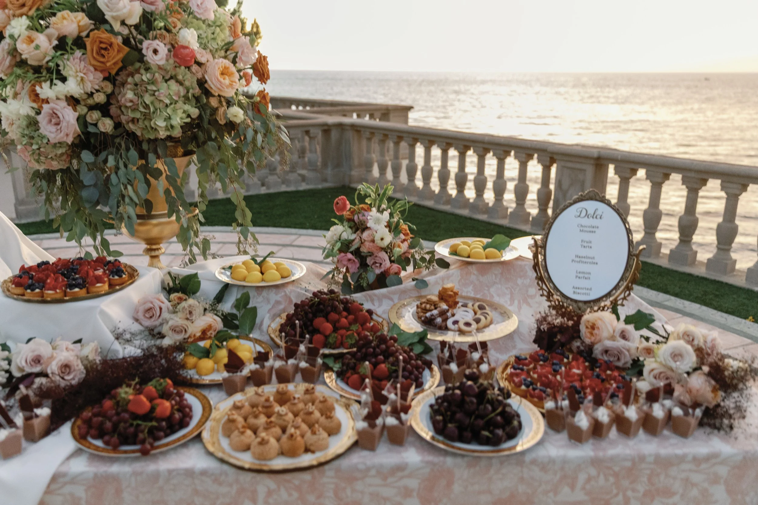 Italian Dessert table served on ornate gold platters and mirror with a printed menu with tiramisu, profiteroles, fruit tarts, cannoli, profiteroles, biscotti, and fresh fruit