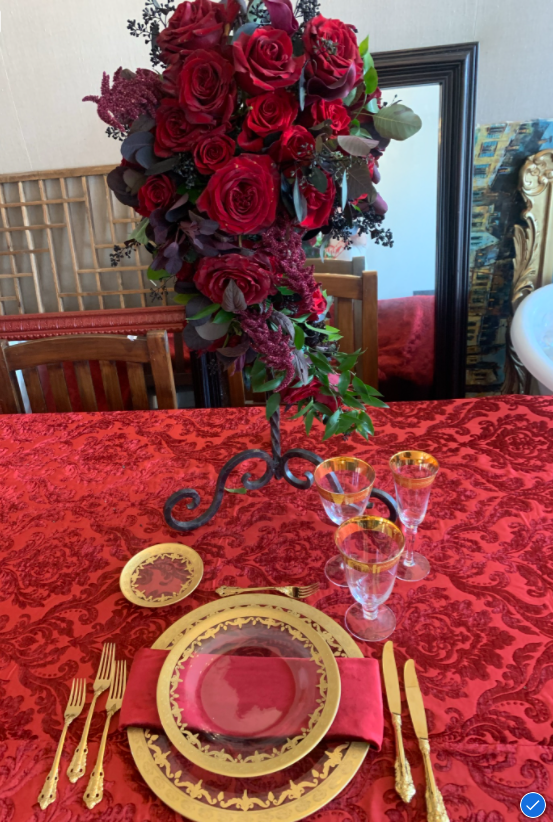 A tall centerpiece of deep red roses and trailing burgundy foliage on a wrought iron stand above a red damask linen. A place setting contains glass chargers with gold rims, gold flatware, gold rimmed glasses, and a red napkin.
