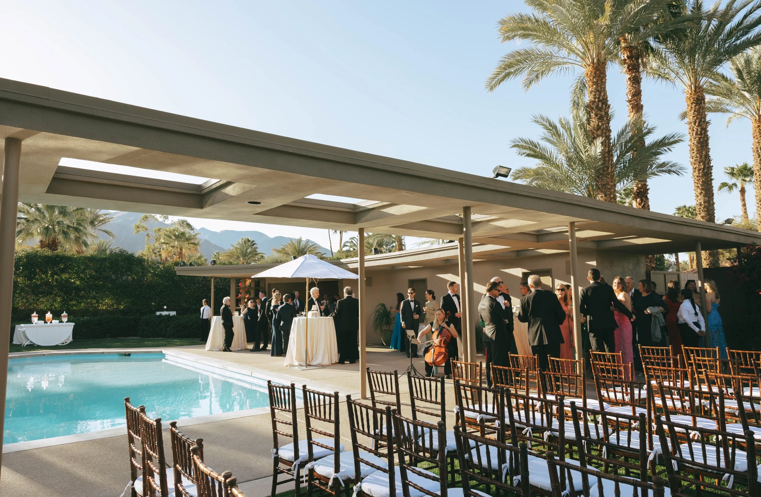 Guests mingle poolside at Twin Palms Estate under the mid-century covered patio, with ceremony chairs and palm trees beyond