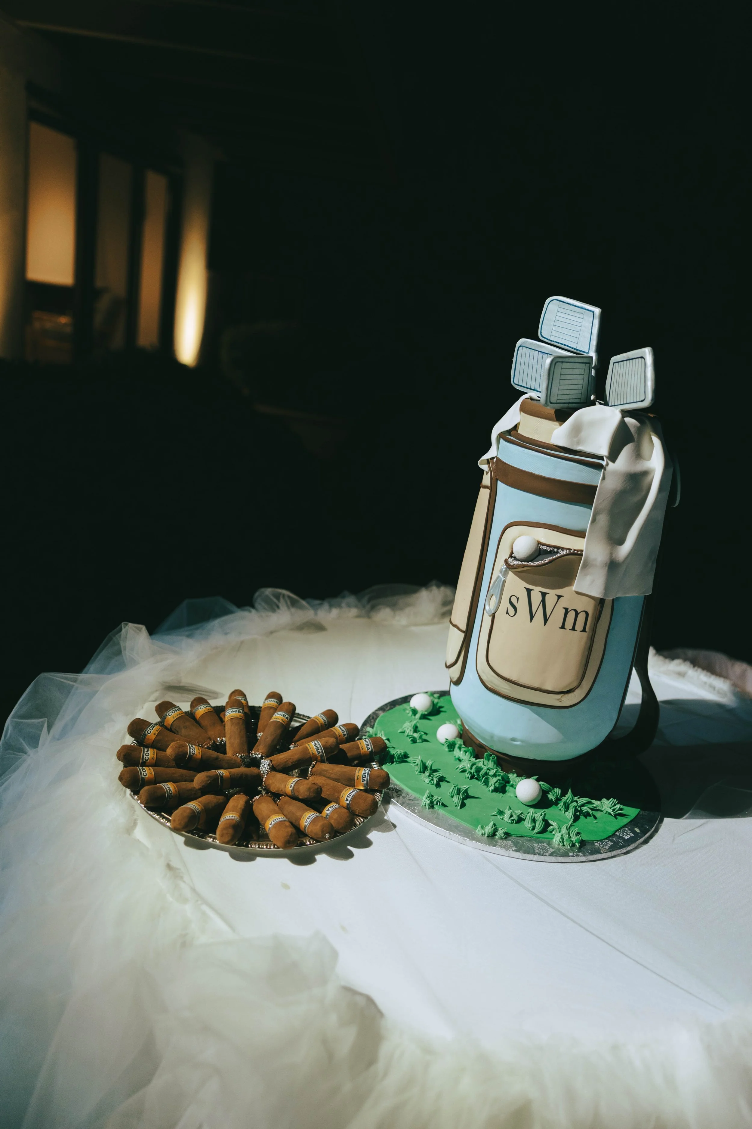 Groom's cake shaped like a monogrammed golf bag, set on a tulle-draped table beside a tray of chocolate cigars