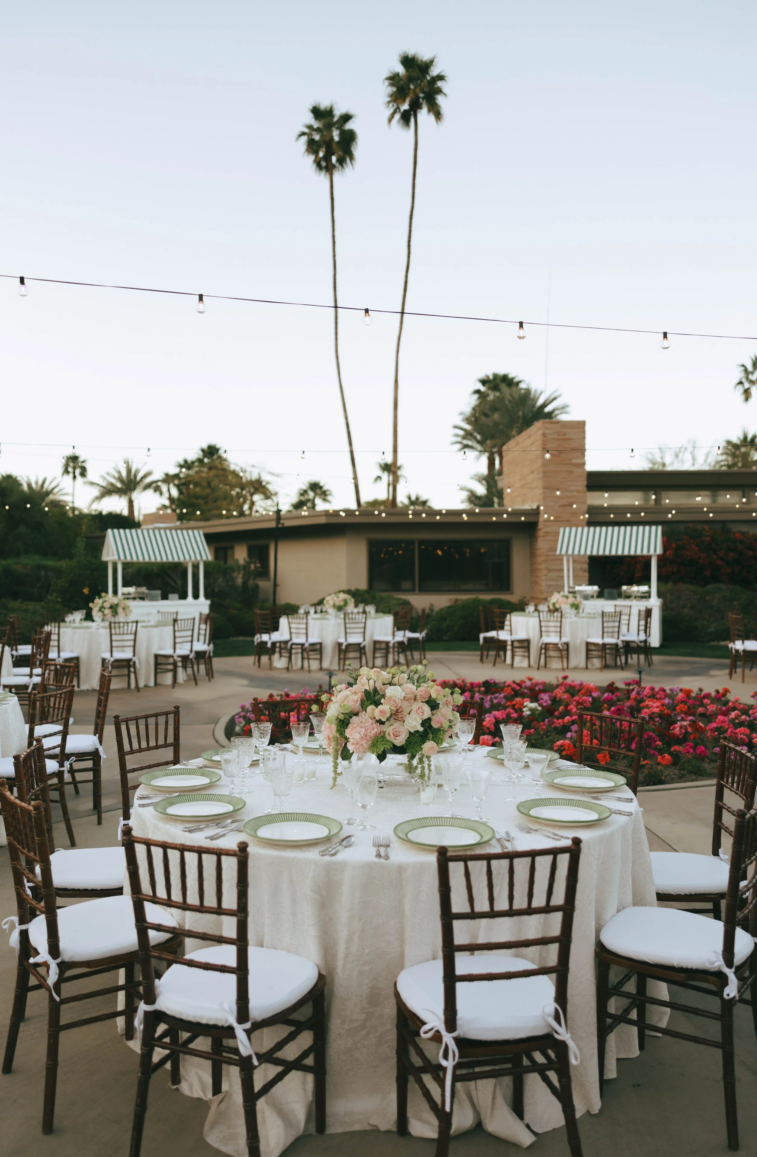 Round reception tables with ivory linens and low blush centerpieces set outdoors beneath palm trees at dusk