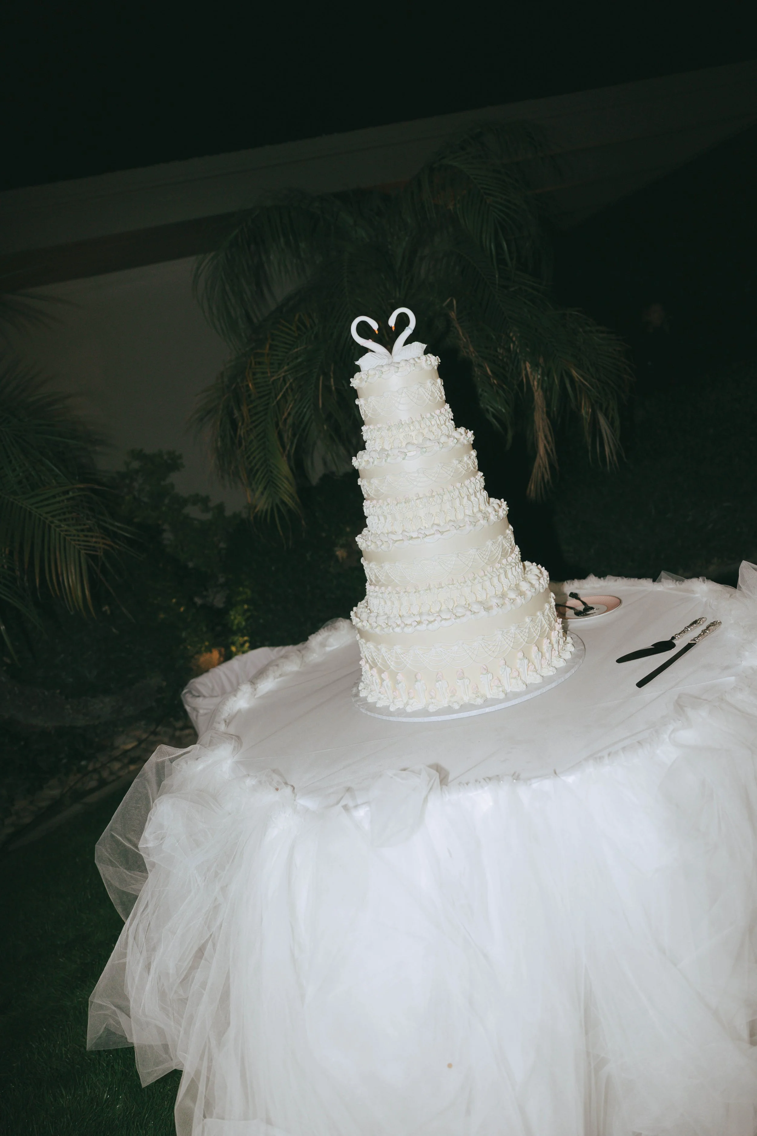 A white multitierd wedding cake on a a white table cloth surrounded with tulle. Two swans making a heart top the cake.