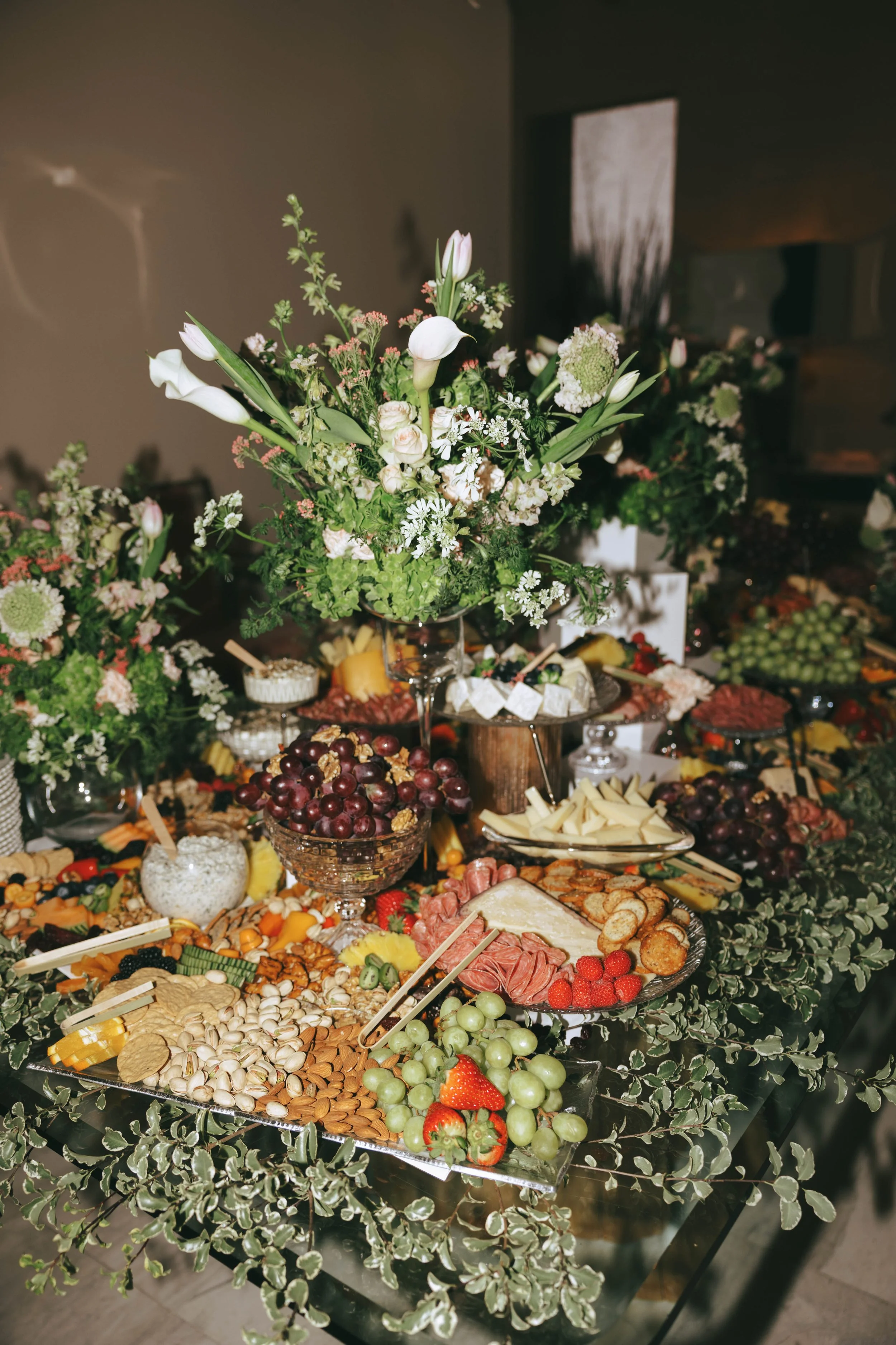 Abundant charcuterie display of cured meats, cheeses, nuts and fruit beneath a tall white and green floral arrangement