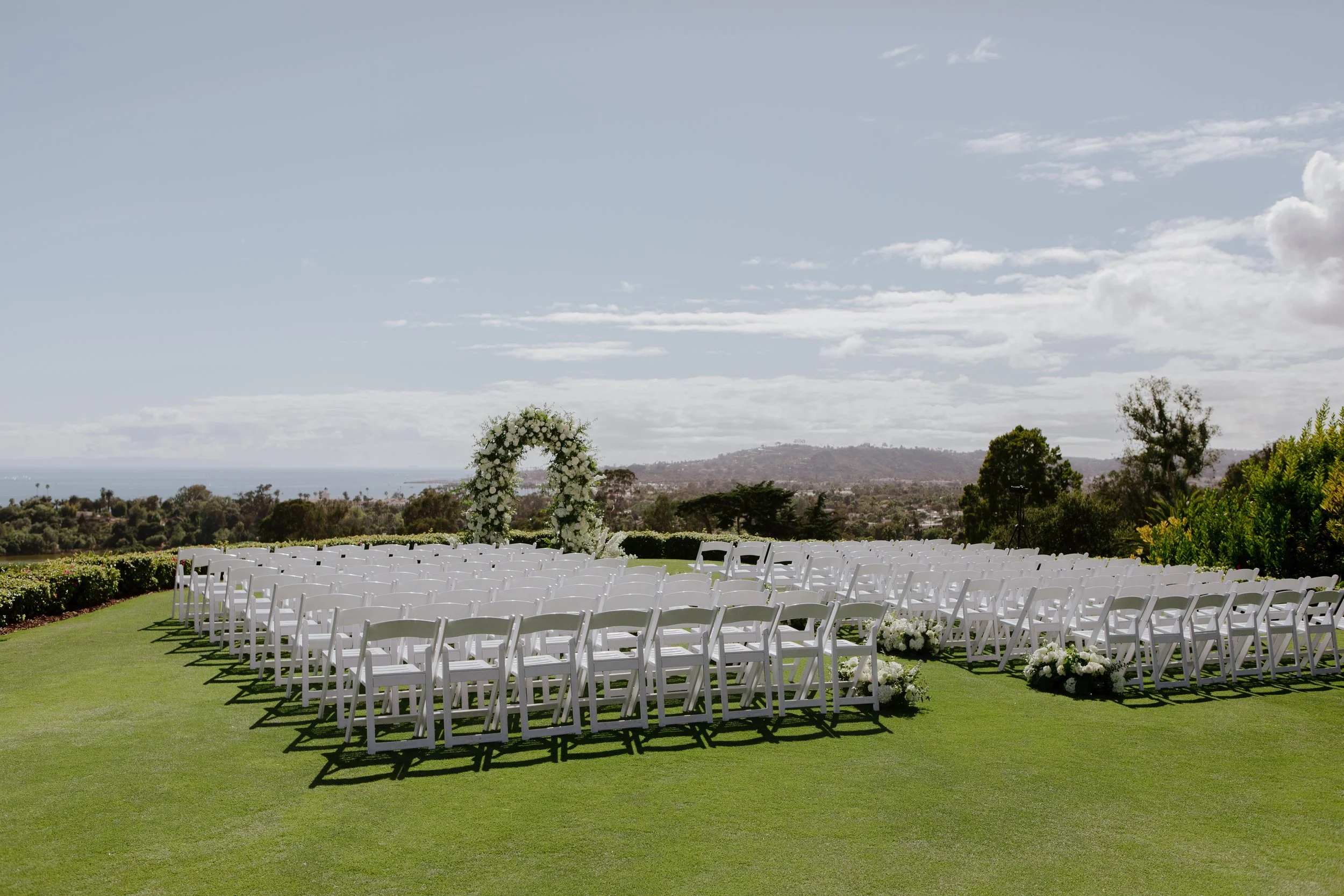 Outdoor ceremony set on the lawn, rows of white chairs facing a white floral arch with the ocean beyond