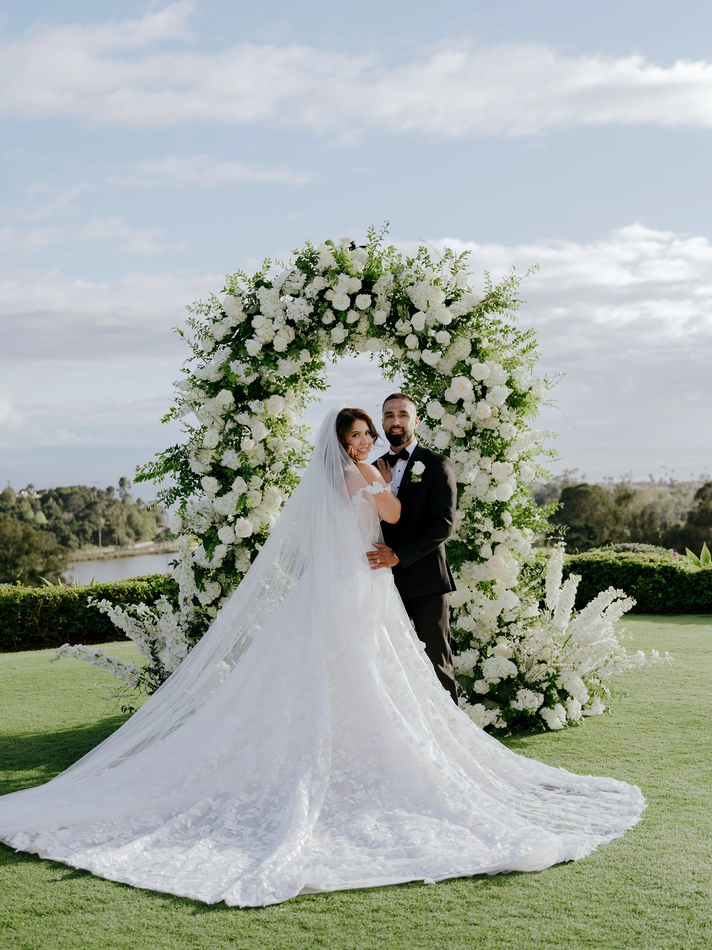 Bride and groom beneath the white rose and greenery arch, her Galia Lahav gown and cathedral veil spread across the grass
