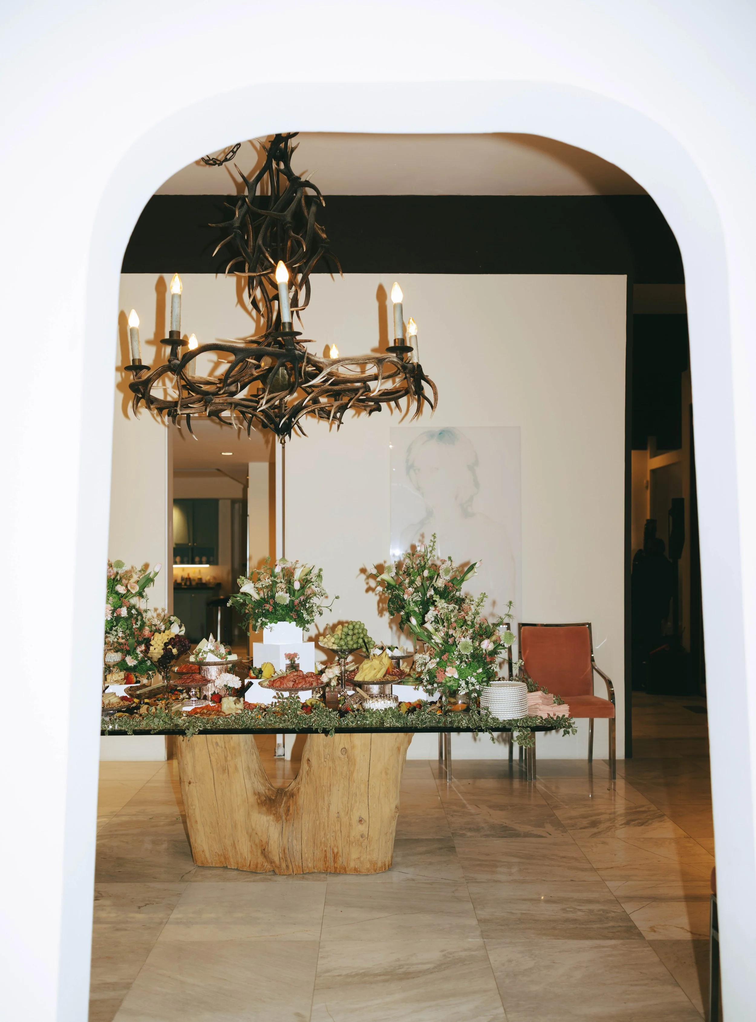 View through an arched doorway to a charcuterie spread on a wood table beneath an antler chandelier