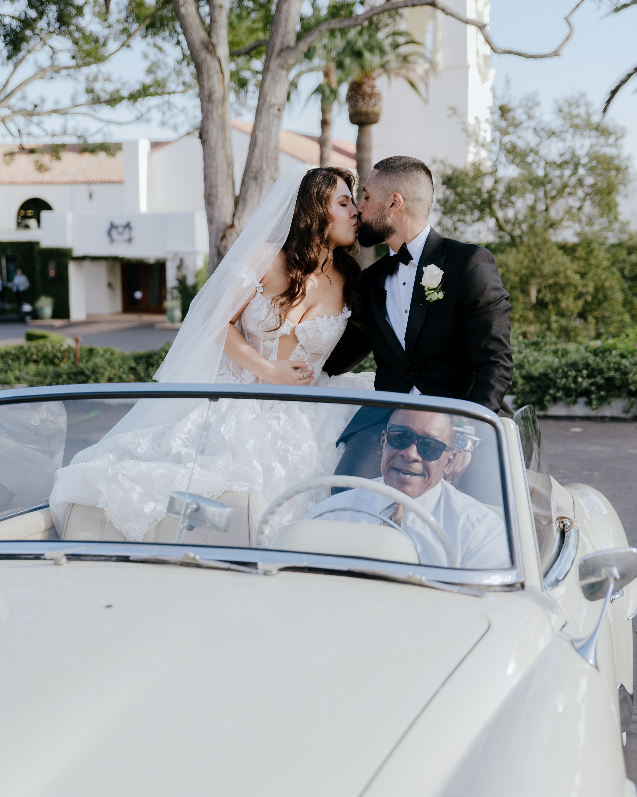 Bride and Groom kiss in the back of their white vintage convertible getaway car. Palm Trees and the Montecito Club in the background.