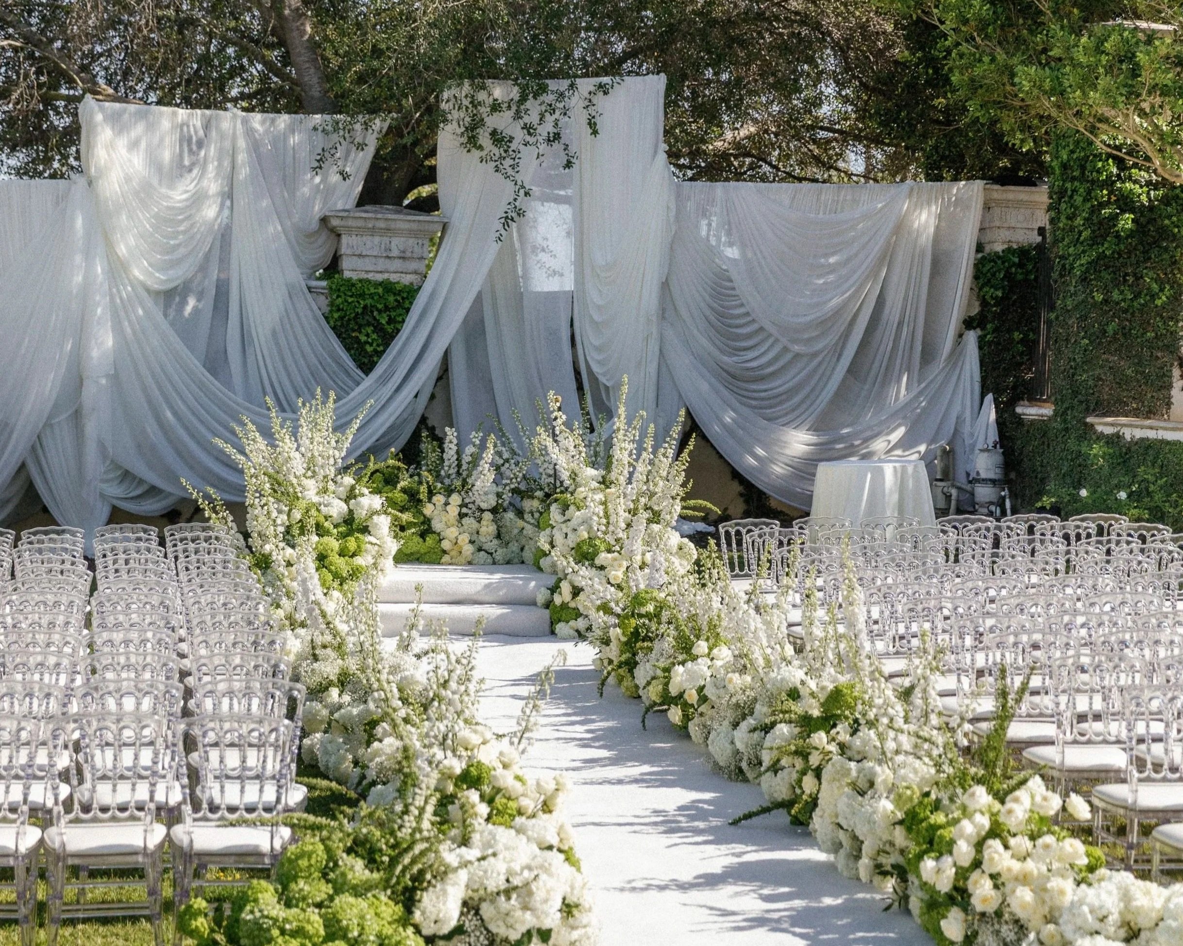 White and green garden ceremony with ghost chairs and a draped altar at The Colony Estate, Camarillo
