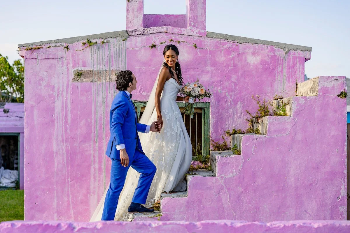 Black bride in a white Wedding dress and white groom in a blue suit holding hands and walking up the stairs of a historic pink building on a tropical island, showcasing a diverse destination wedding by Center of Attention Events in the Bahamas.