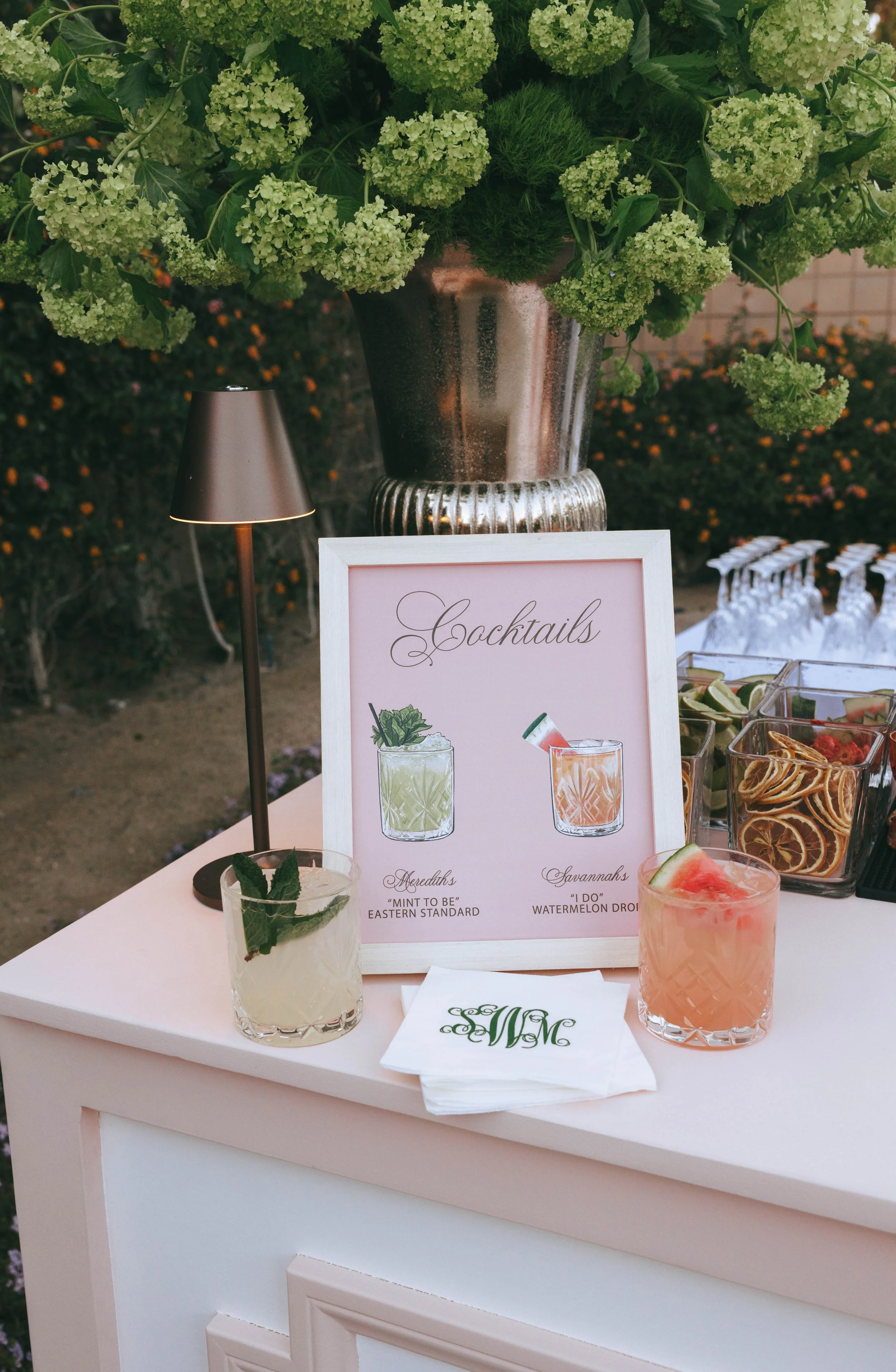 Blush bar with a framed "Cocktails" sign illustrating the couple's two signature drinks, beside monogrammed napkins