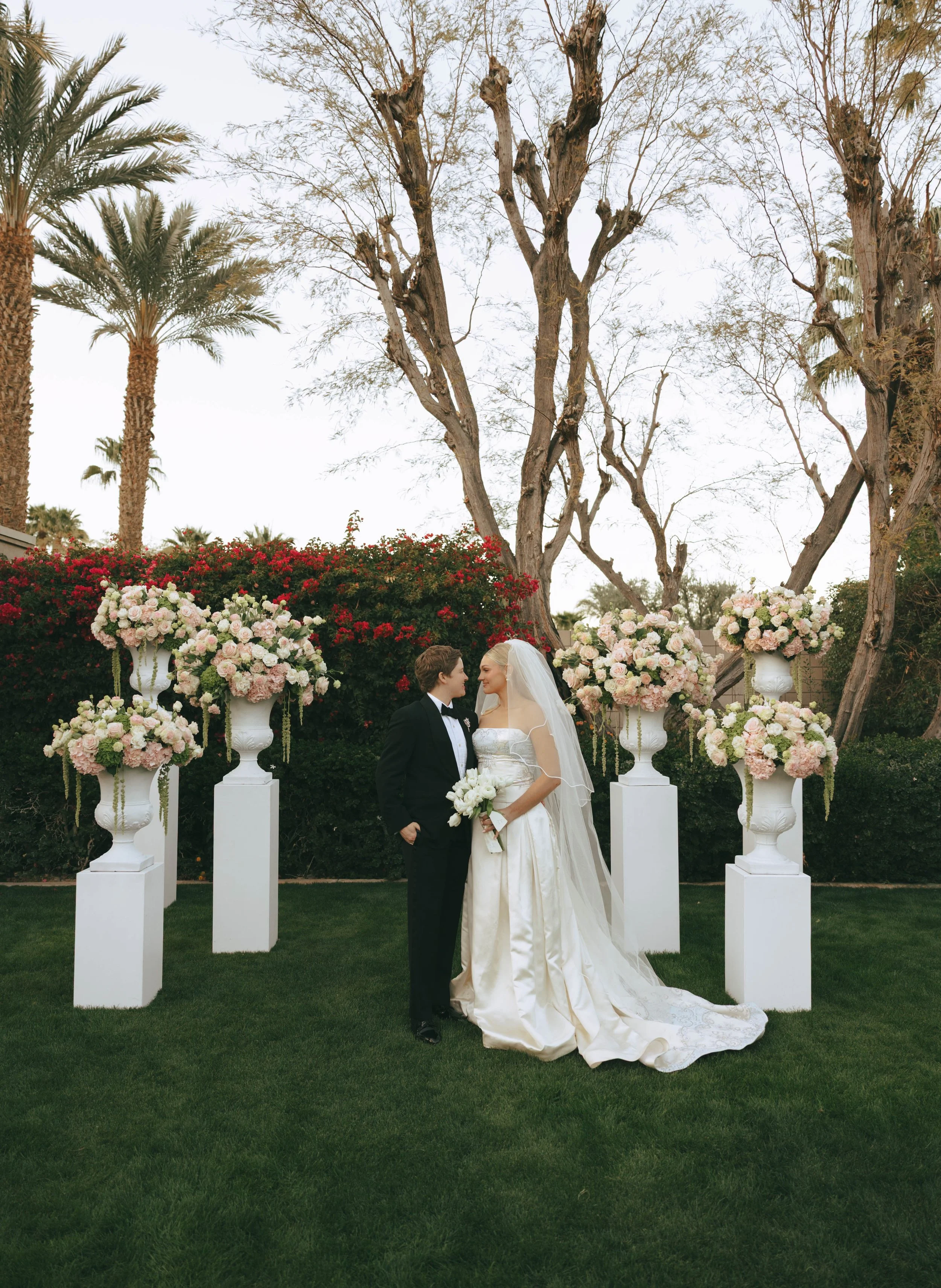 Bride holding a white bouquet and groom posing for photos around white, pink, and green floral arrangements on pedestals. A green hedge with red flowers fills the background.