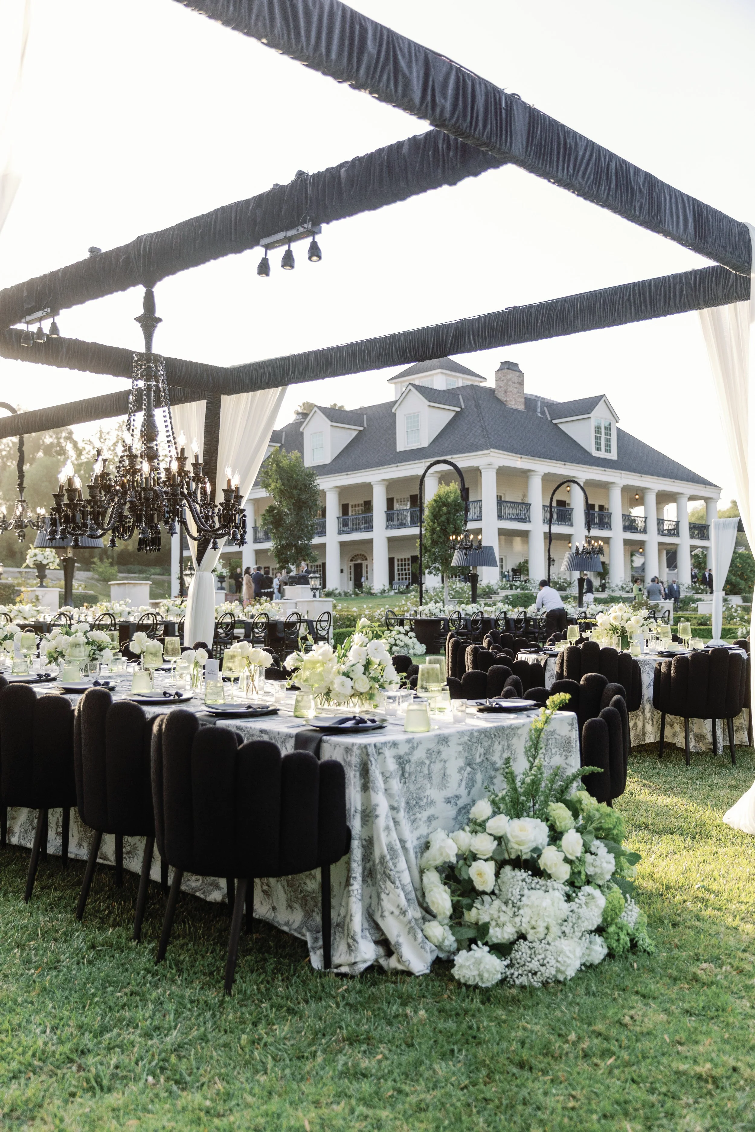 Wedding reception tables beneath a black cabana with white draping and a black chandelier. Black fluffy chairs along the edge of the table. White main house of the Colony Estate in the background.
