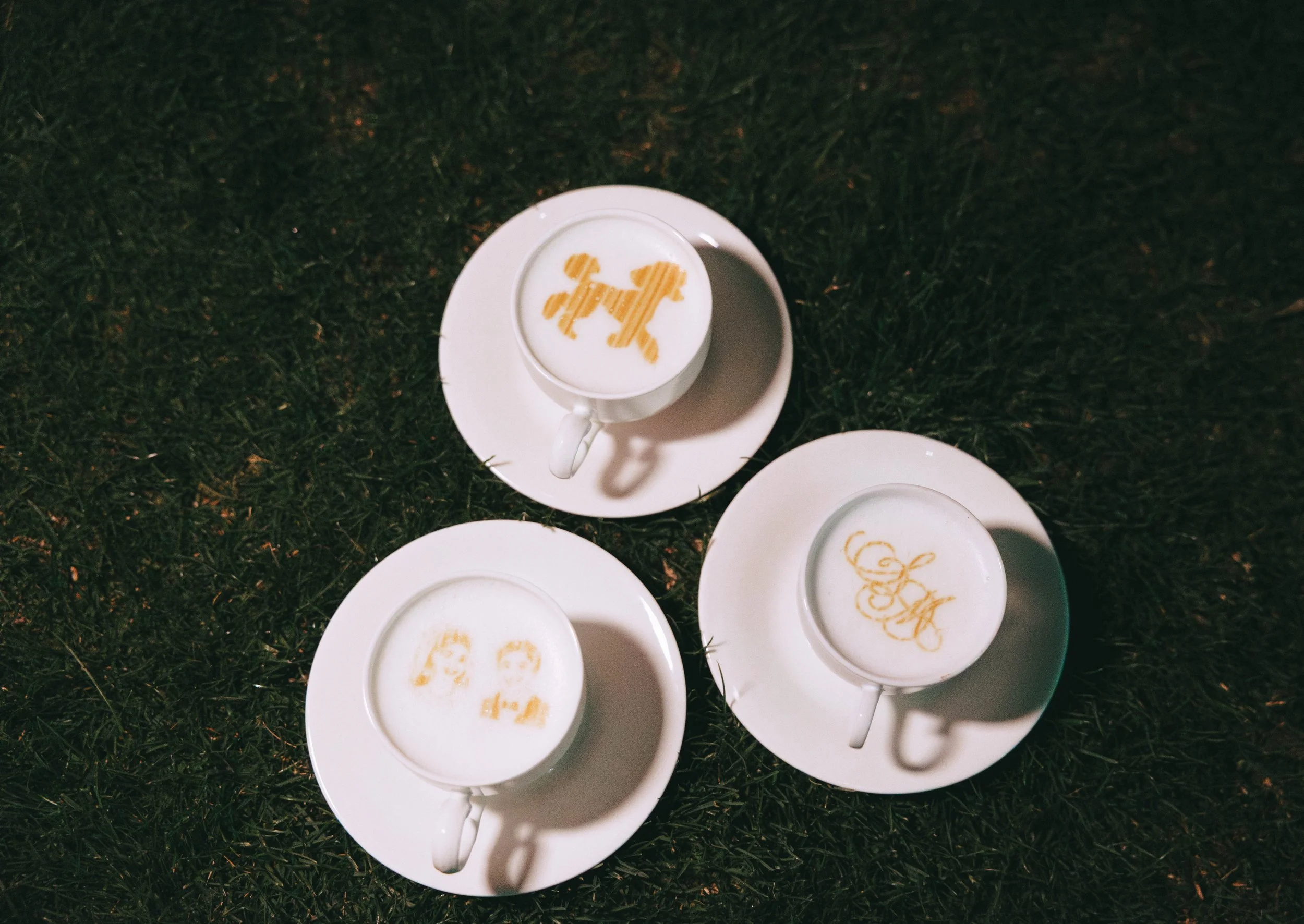 Three cappuccinos on white saucers in the grass, each finished with monogram and cartoon-portrait latte art