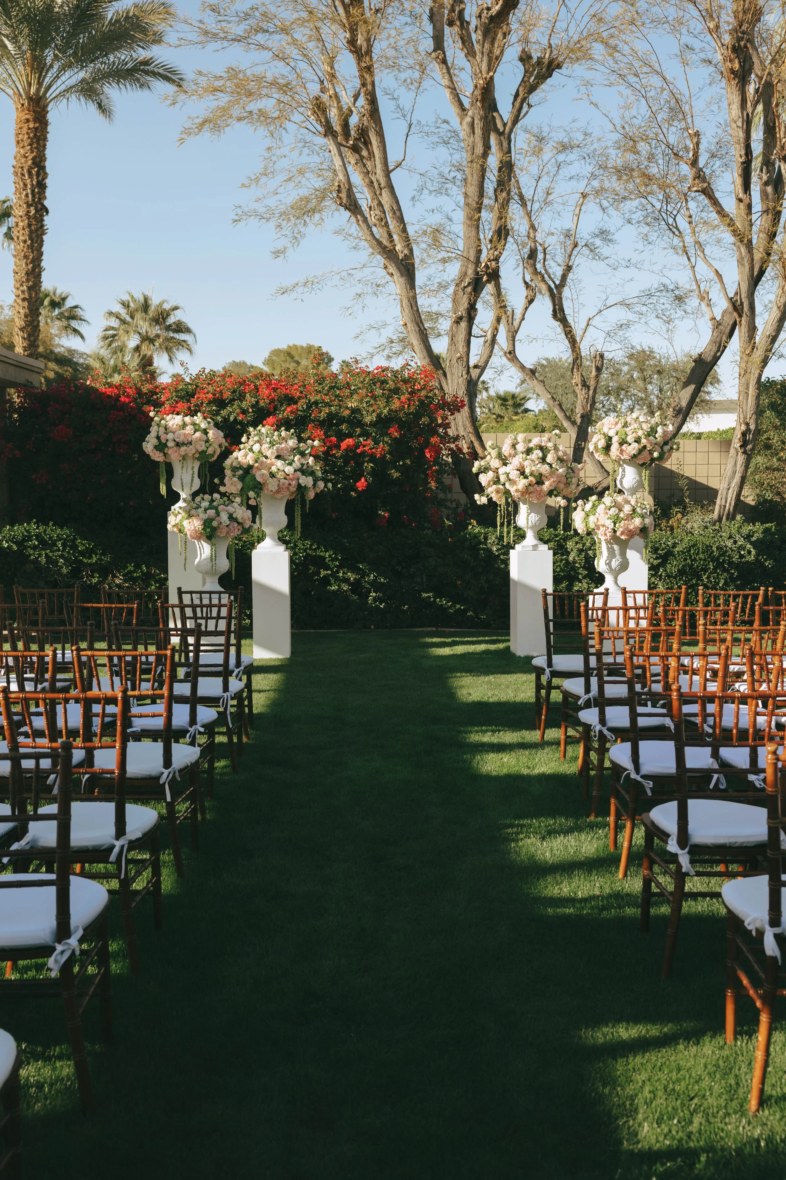 Ceremony aisle on the lawn lined with wooden chairs, leading to white pedestals with blush floral urns and bougainvillea