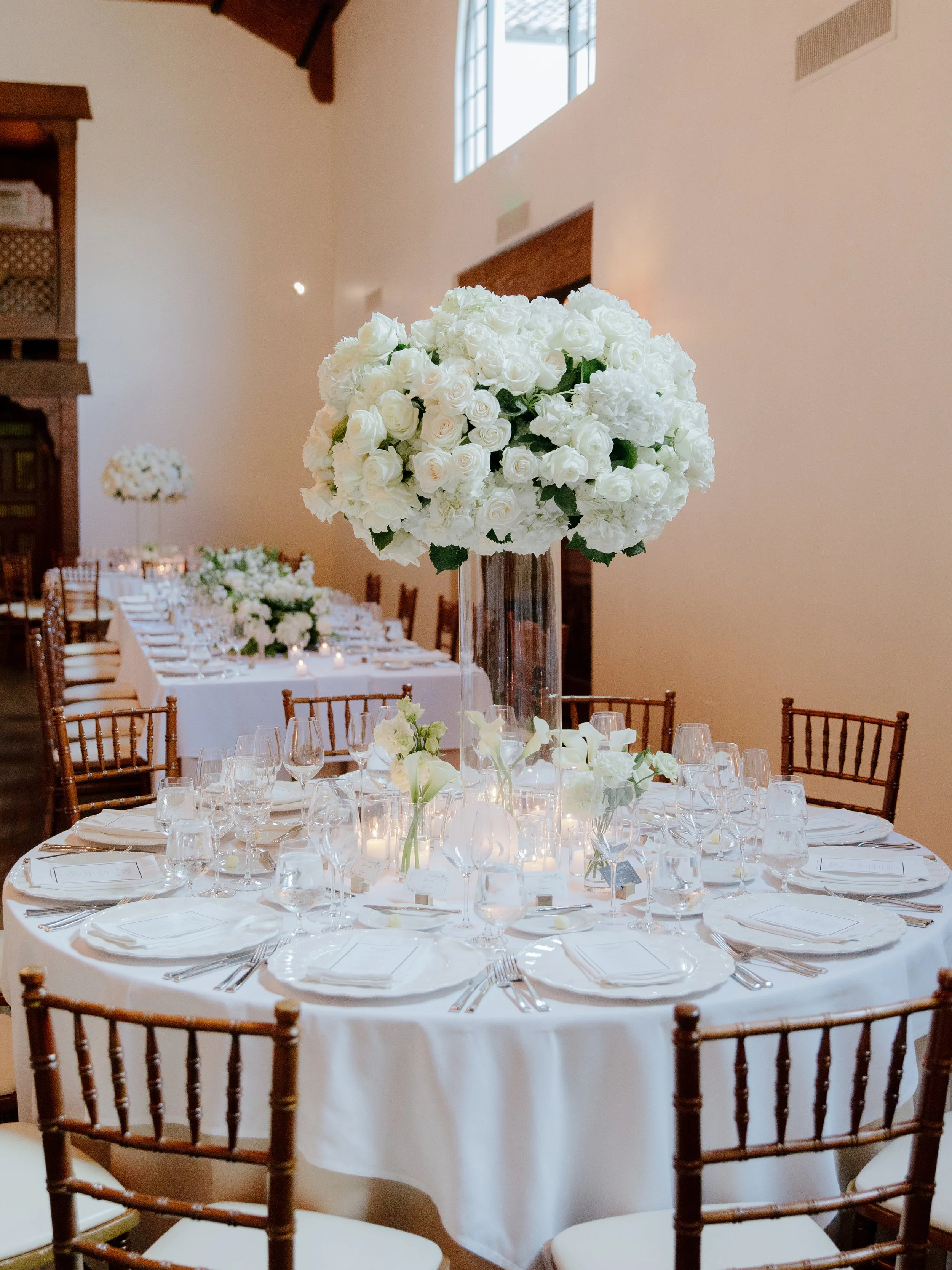 Round reception table with a tall white hydrangea and rose centerpiece, gold chiavari chairs and candlelight