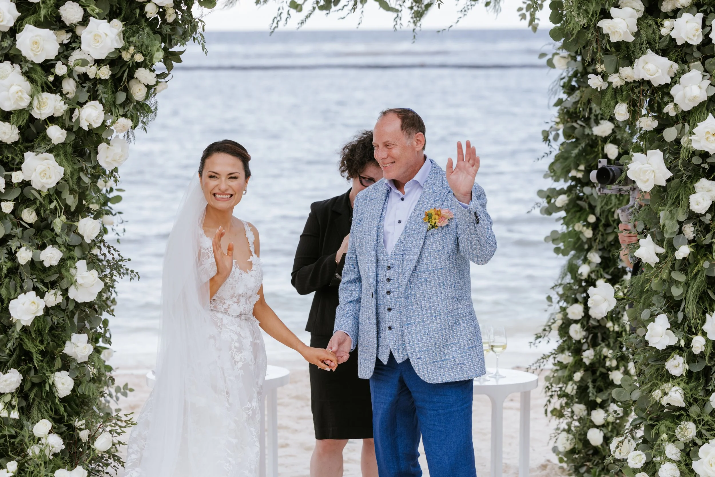 Bride and groom at the beachfront altar framed by a white rose and greenery arch, the officiant and ocean behind them