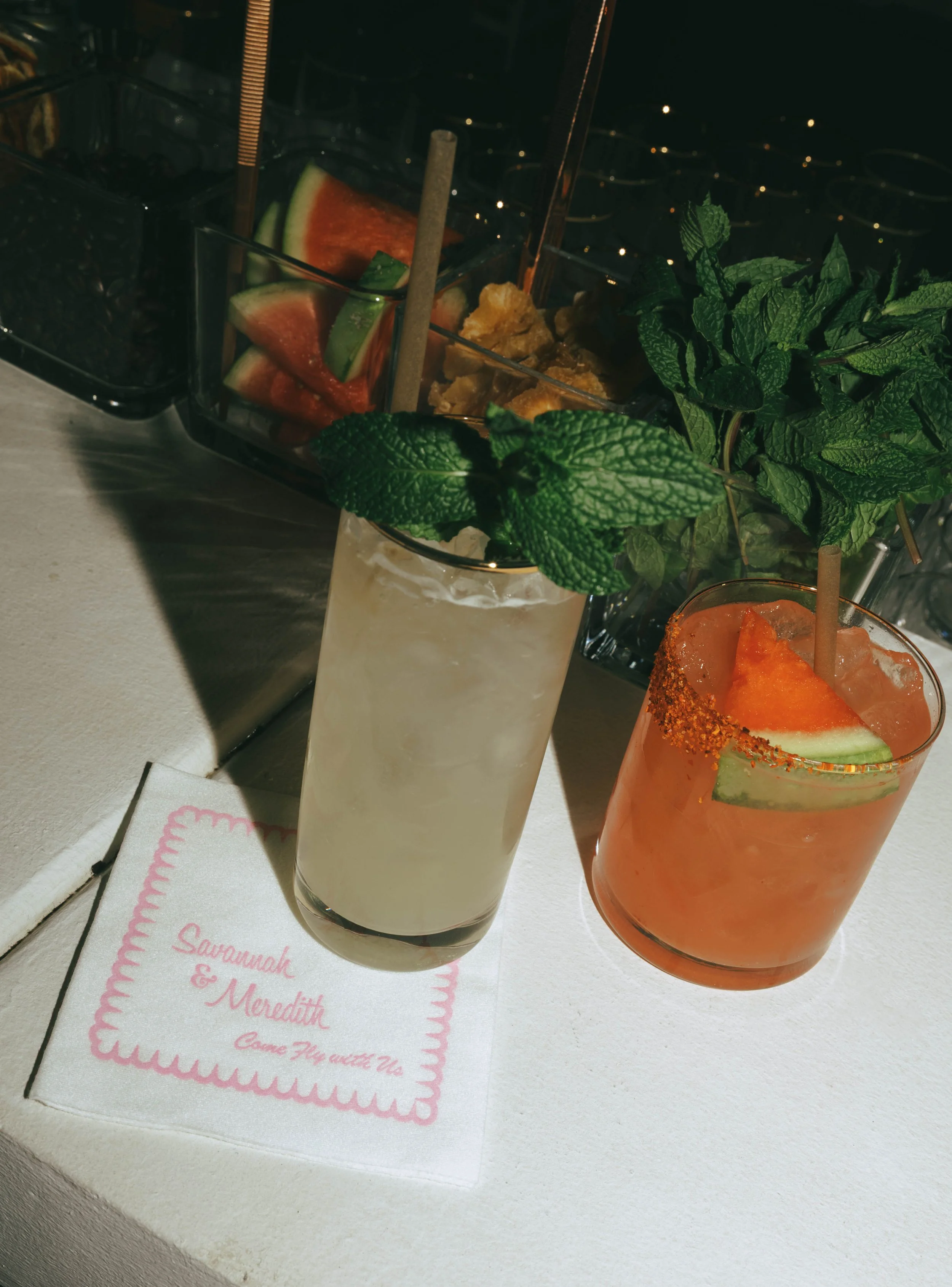 Two signature cocktails garnished with mint and watermelon on a bar ledge beside a pink monogrammed cocktail napkin