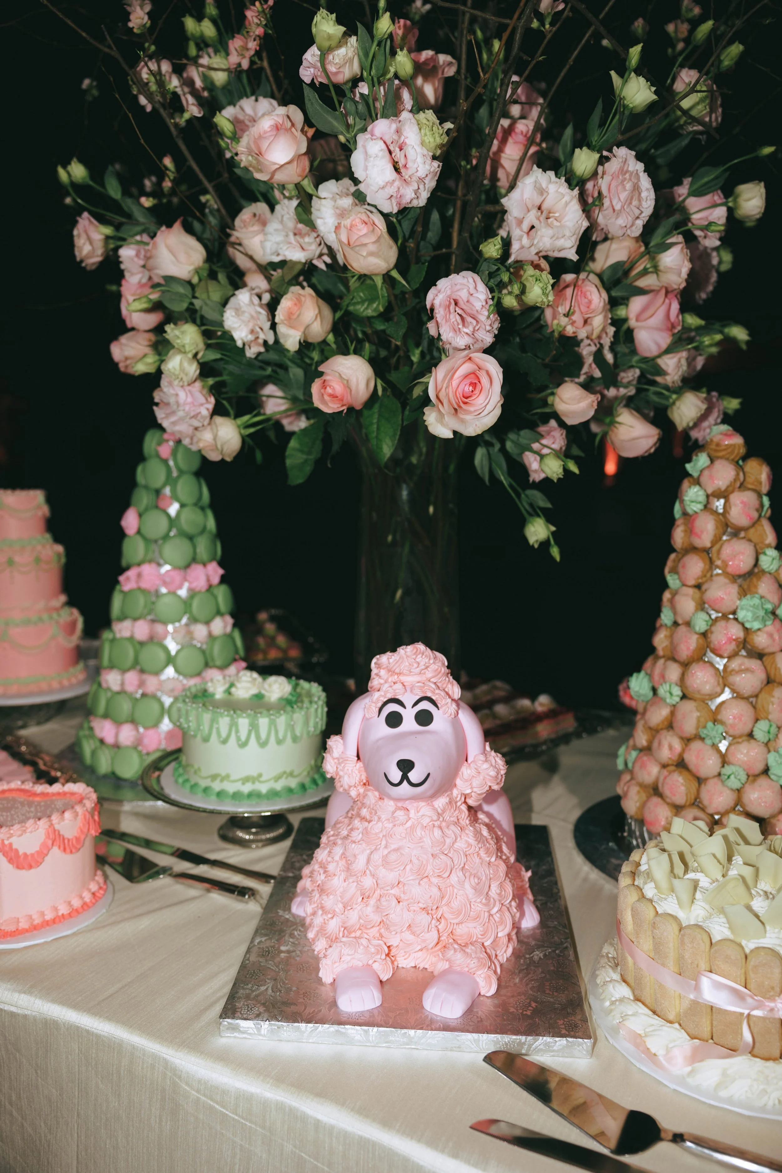 Welcome party dessert table with a pink poodle-shaped cake, pastel layer cakes, croquembouches and towering pink roses