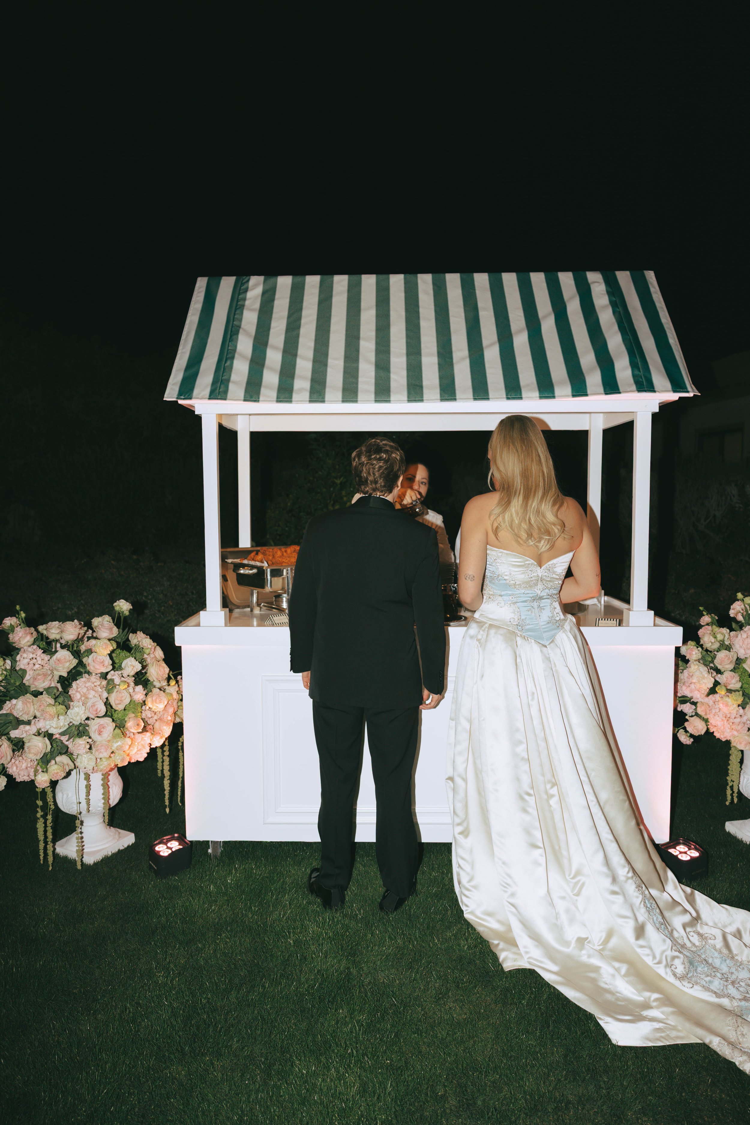 Bride and groom at a green-and-white striped bar at night, surrounded by blush floral arrangements