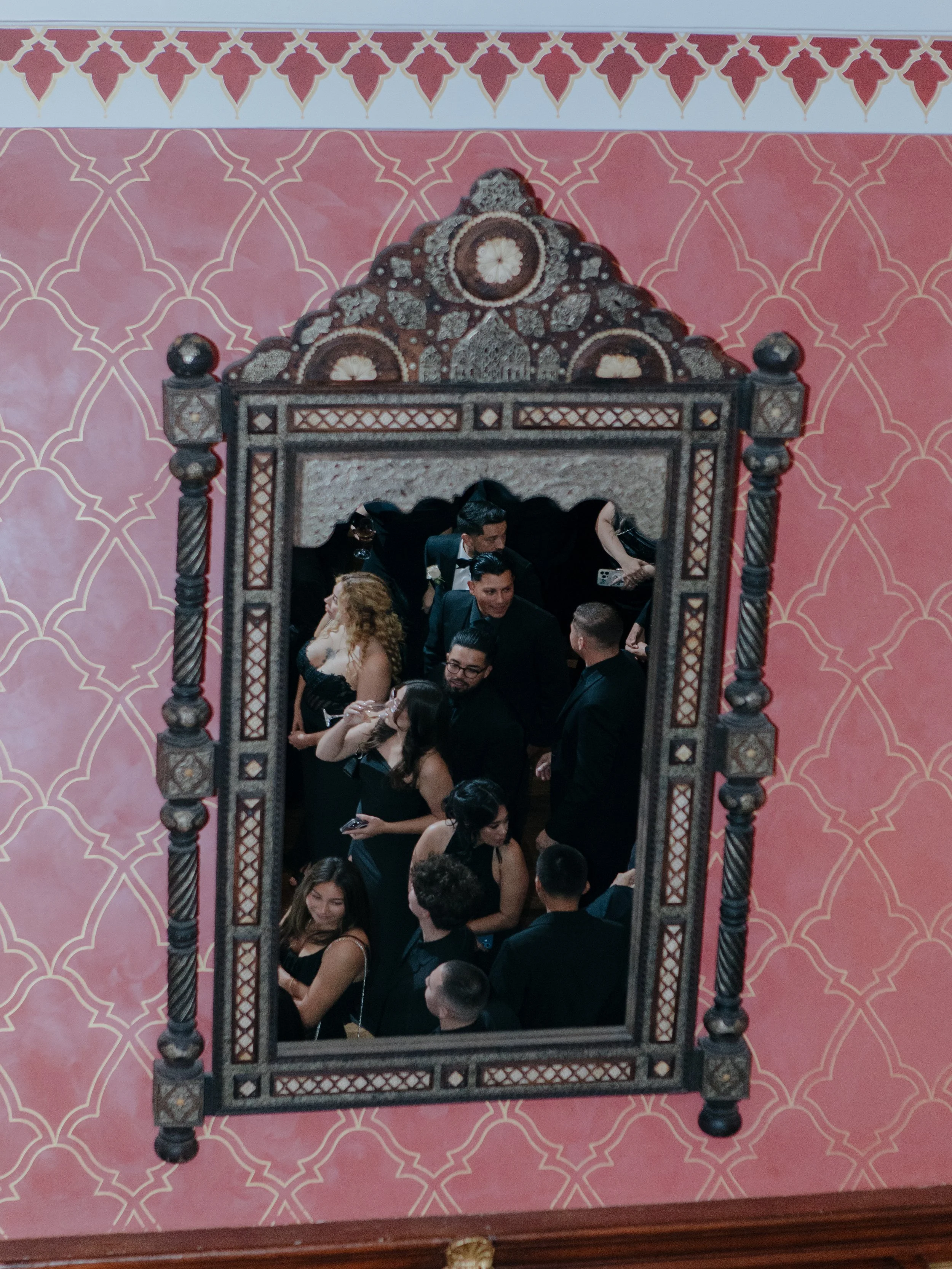 Guests mingling reflected in an ornate mirror on the Montecito Club's pink patterned wall