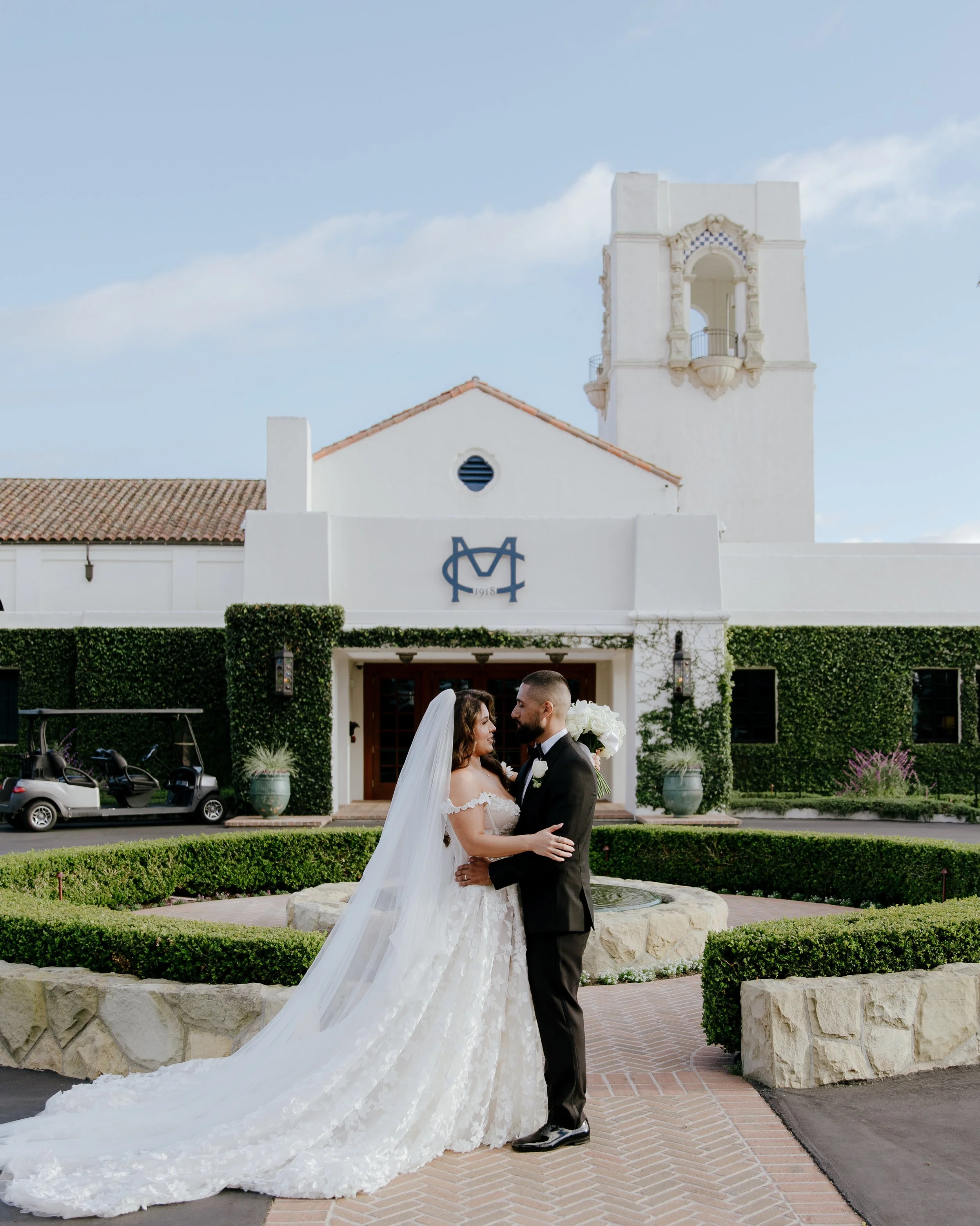 Bride and Groom hold each other in front of the hedge-encircled fountain of the Montecito Club clubhouse, the ivy-covered building's bell tower, and monogram behind them
