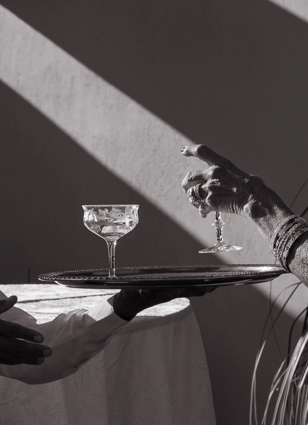 A black and white photo of a hand grabbing a drink in a crystal glass off a silver tray in a waiters hand.