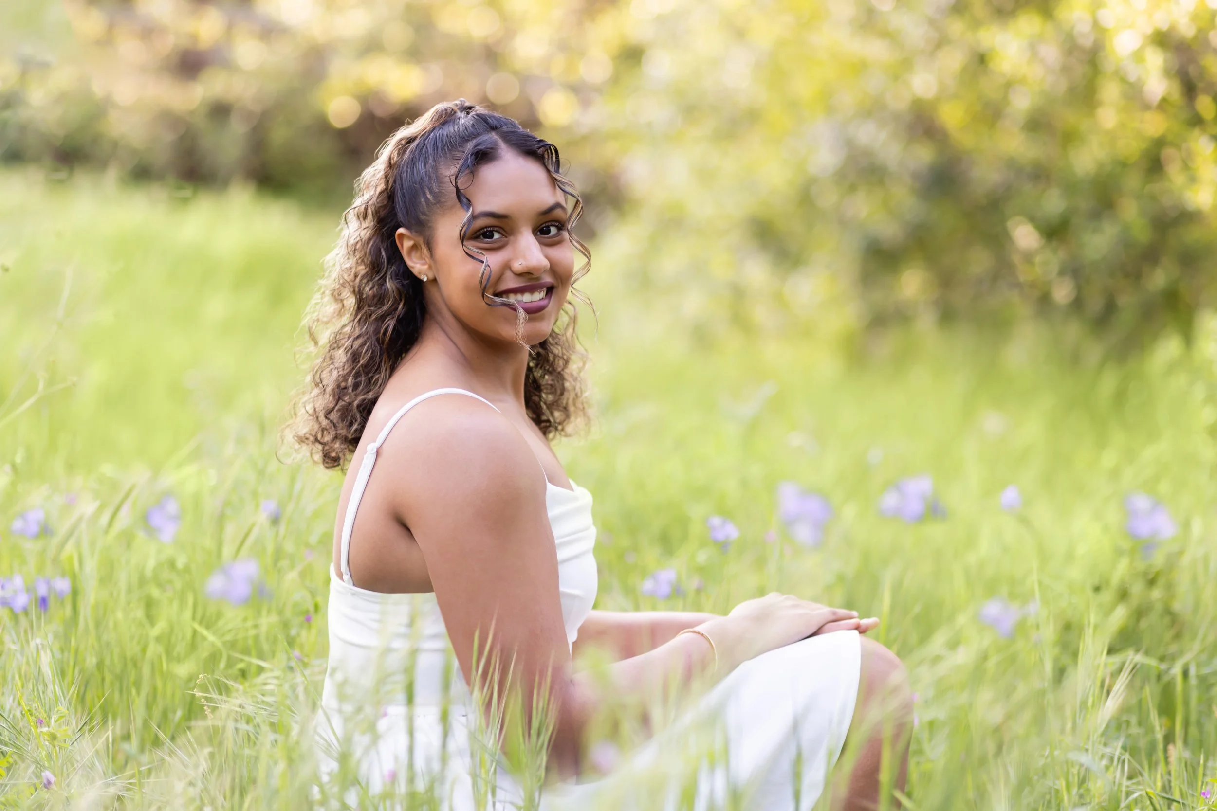 high school senior girl sits in a grass field with purple flowers around her at veterans memorial park in Roseville, california